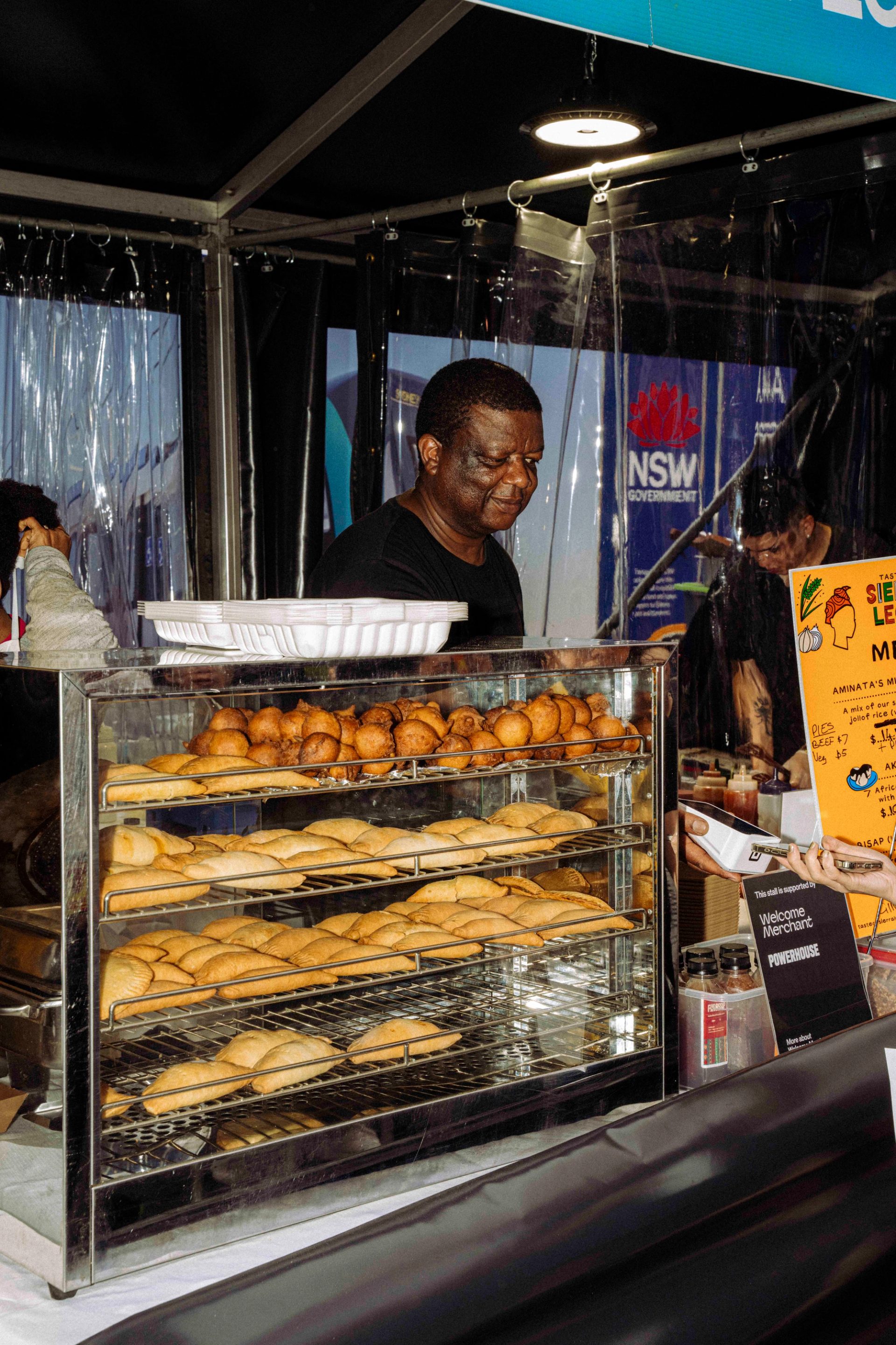 A vendor with short hair and wearing a black top stands behind a glass display filled with four neat rows of pastries and fritters at a busy food stall.