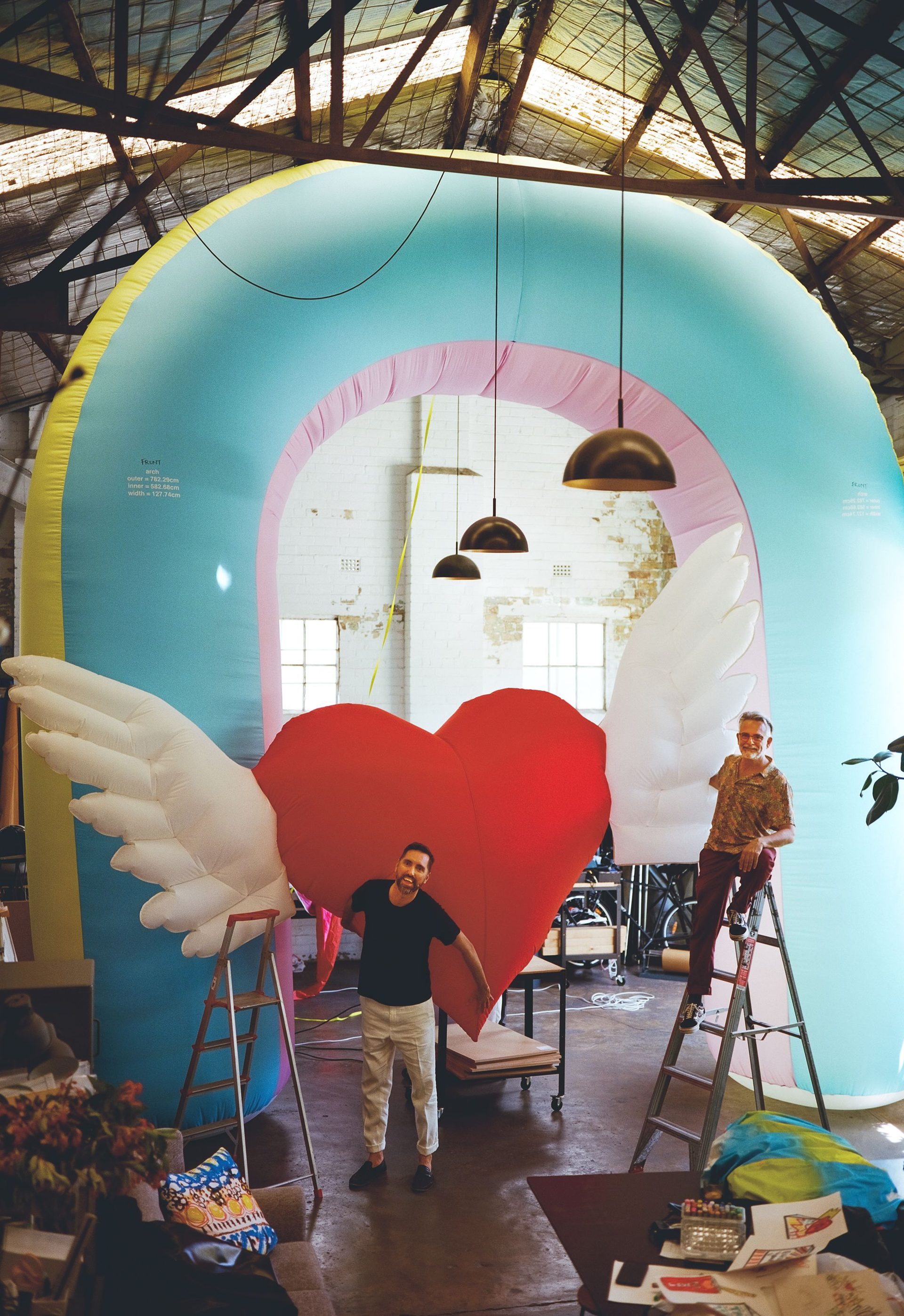 Two men in an art studio. One man rests halfway up a ladder and the other stands on the ground. His arms reach behind him to hold a large inflatable love heart with wings. There is a large blue, yellow and light pink inflatable arch behind them.