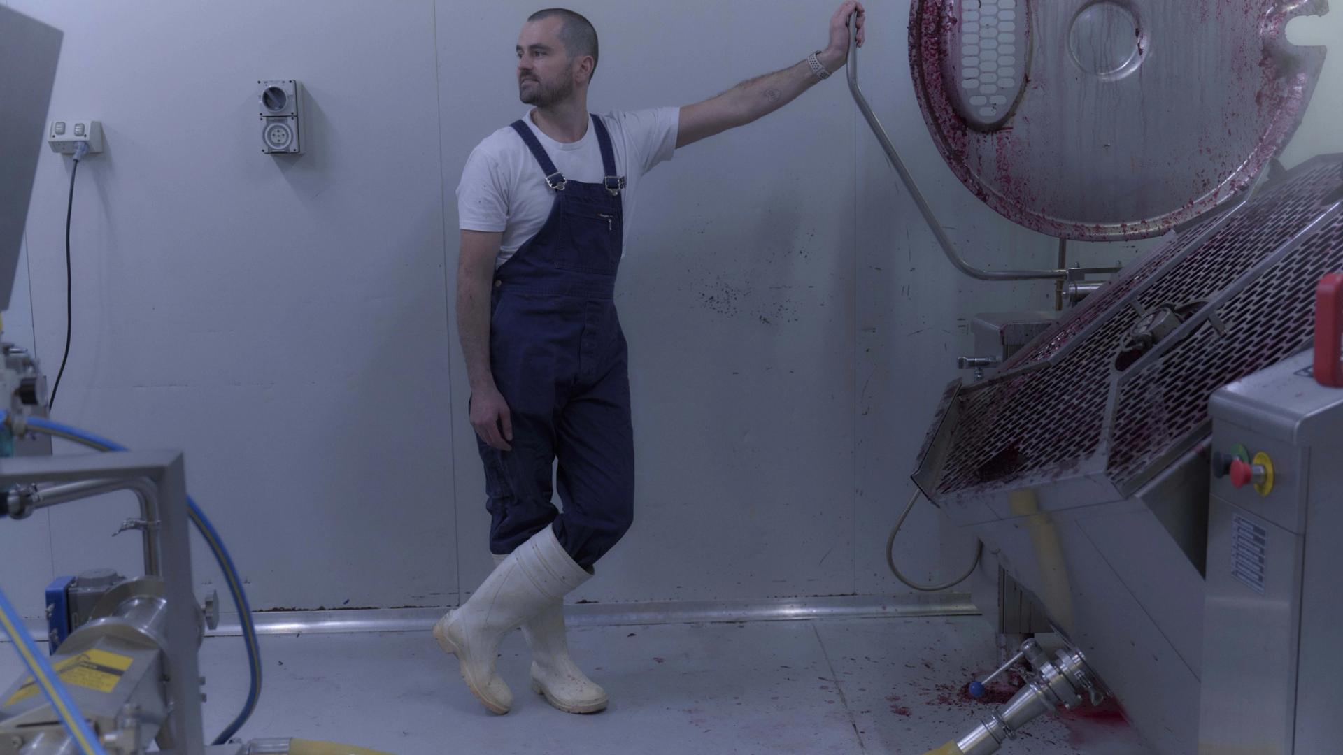 A man dressed in blue overalls, a white t-shirt, and white rubber boots stands in a sterile, industrial room next to a large piece of stainless steel equipment. He leans casually against the machine, which has traces of red liquid, possibly from food processing. The scene is minimalist, with the white walls and floor emphasising the industrial nature of the environment.