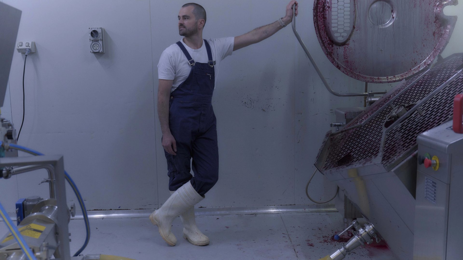 A man dressed in blue overalls, a white t-shirt, and white rubber boots stands in a sterile, industrial room next to a large piece of stainless steel equipment. He leans casually against the machine, which has traces of red liquid, possibly from food processing. The scene is minimalist, with the white walls and floor emphasising the industrial nature of the environment.