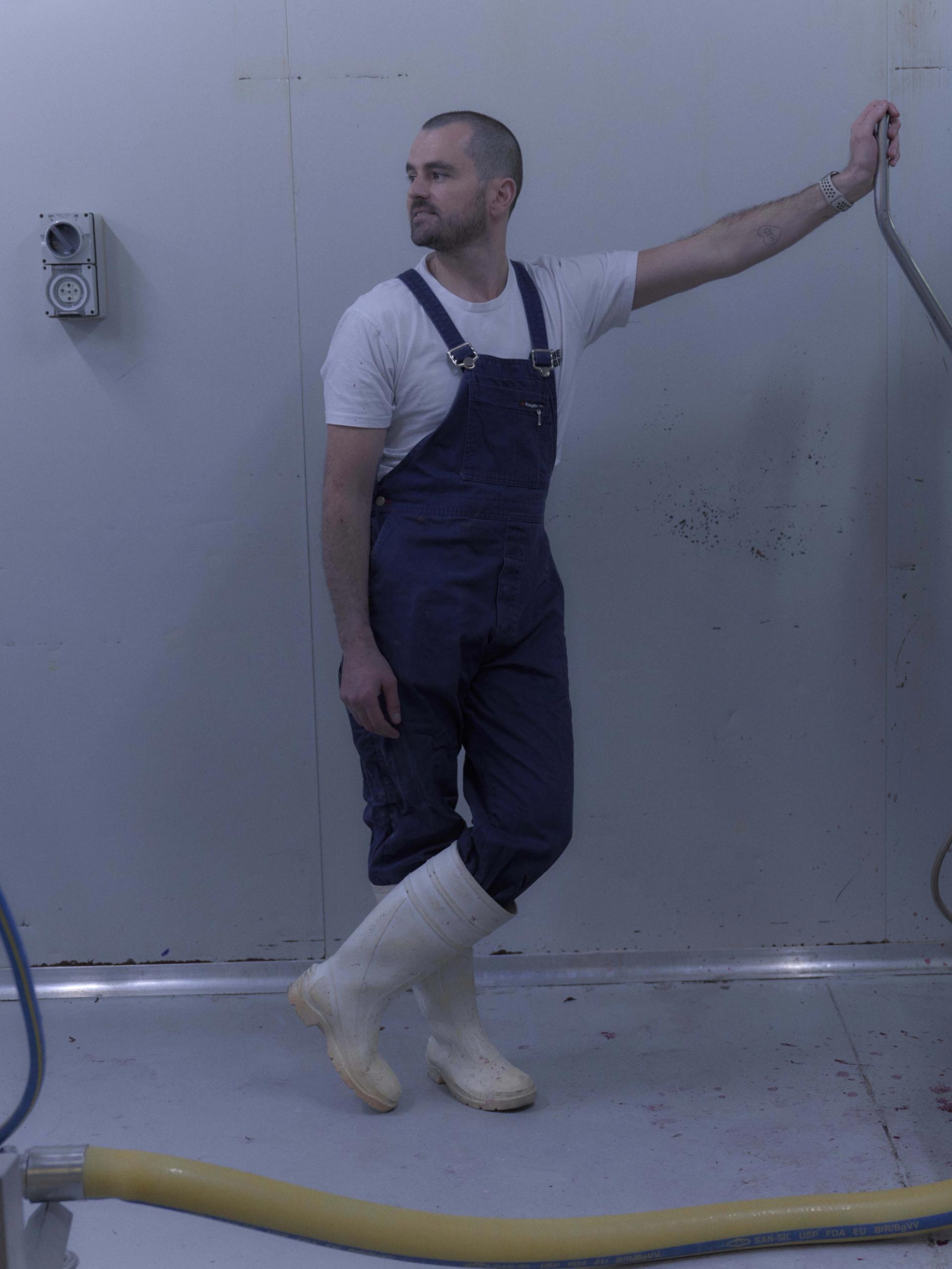 A man dressed in blue overalls, a white t-shirt, and white rubber boots stands in a sterile, industrial room next to a large piece of stainless steel equipment. He leans casually against the machine, which has traces of red liquid, possibly from food processing. The scene is minimalist, with the white walls and floor emphasising the industrial nature of the environment.