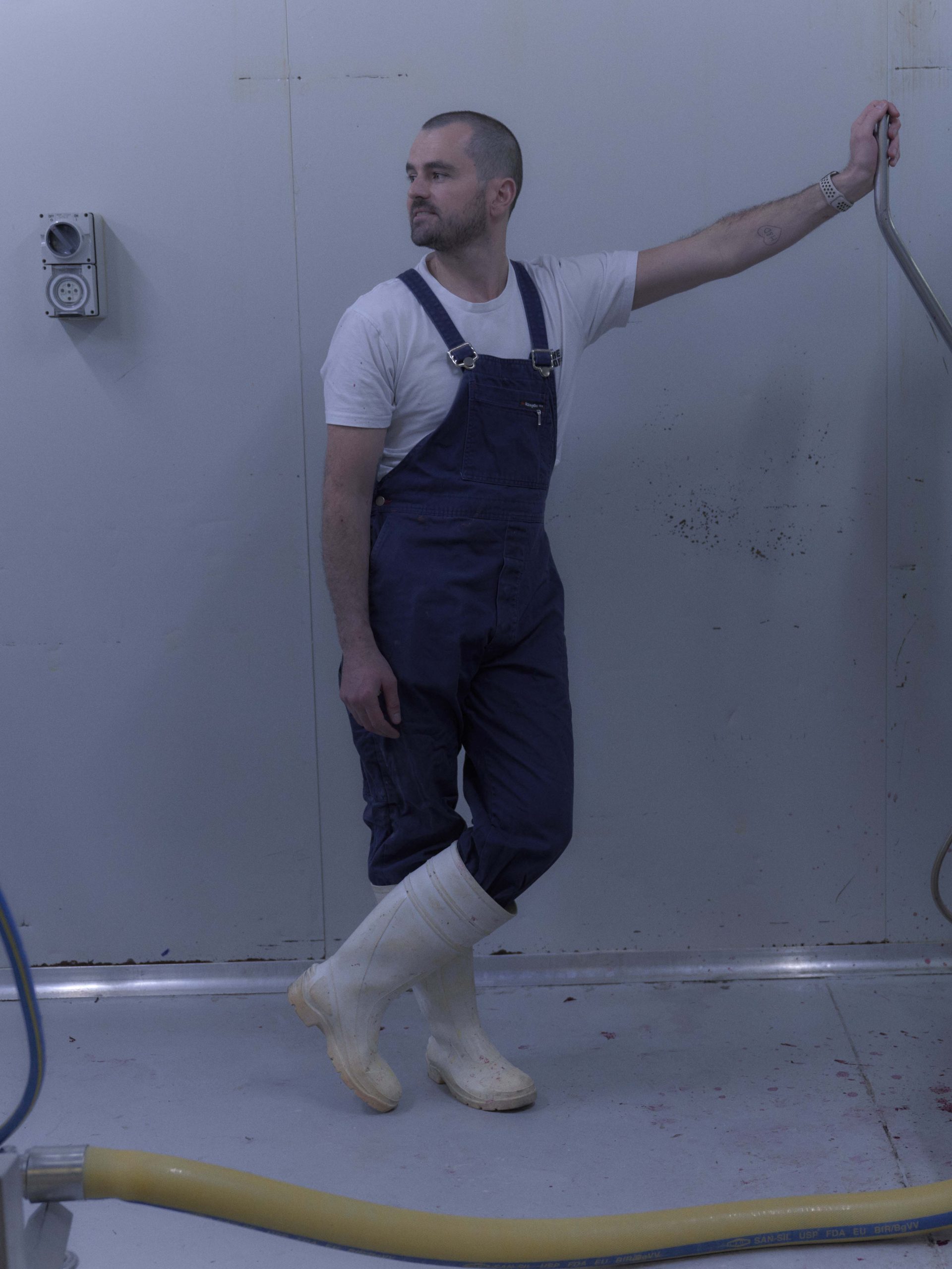 A man dressed in blue overalls, a white t-shirt, and white rubber boots stands in a sterile, industrial room next to a large piece of stainless steel equipment. He leans casually against the machine, which has traces of red liquid, possibly from food processing. The scene is minimalist, with the white walls and floor emphasising the industrial nature of the environment.