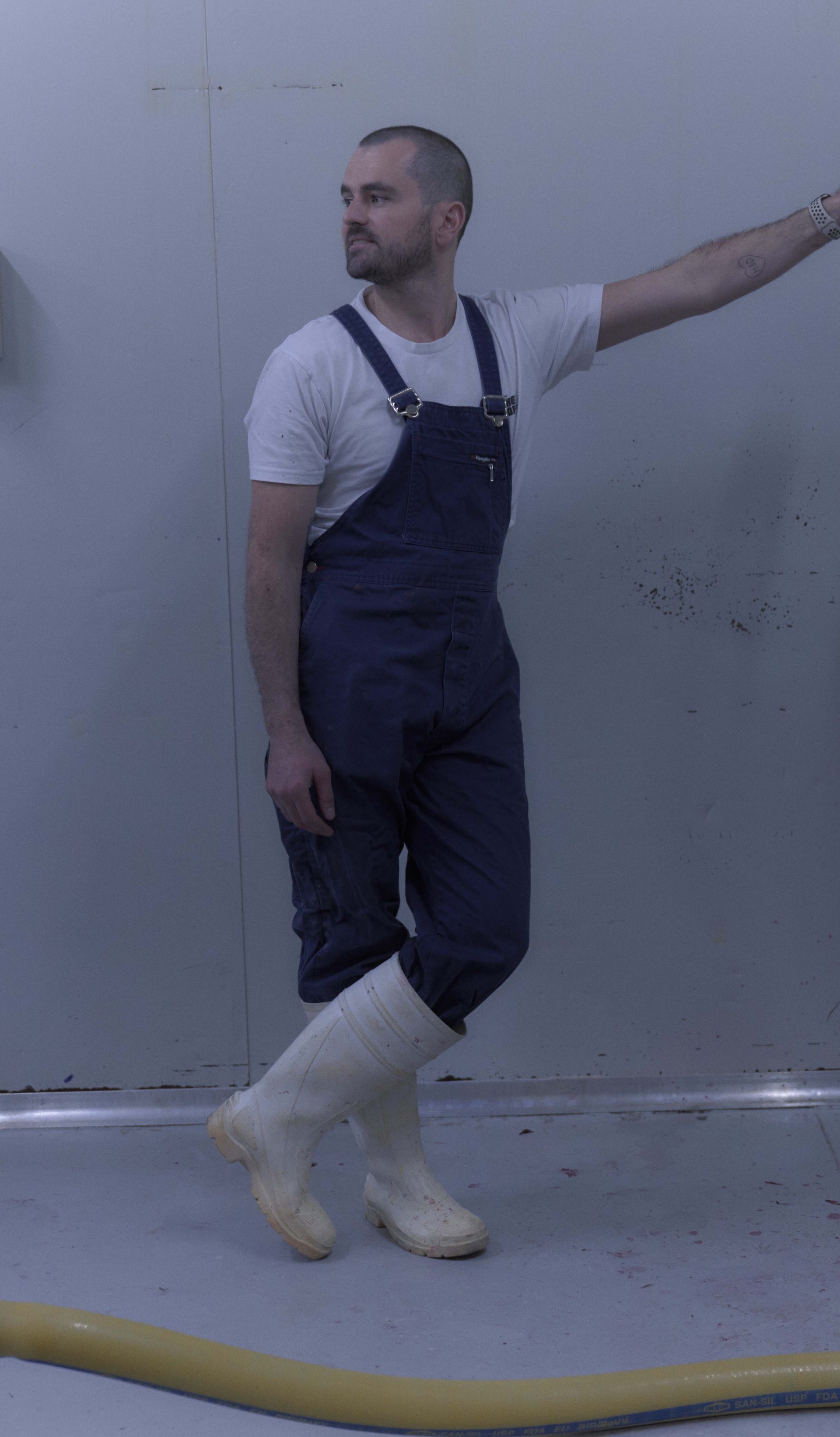 A man dressed in blue overalls, a white t-shirt, and white rubber boots stands in a sterile, industrial room next to a large piece of stainless steel equipment. He leans casually against the machine, which has traces of red liquid, possibly from food processing. The scene is minimalist, with the white walls and floor emphasising the industrial nature of the environment.