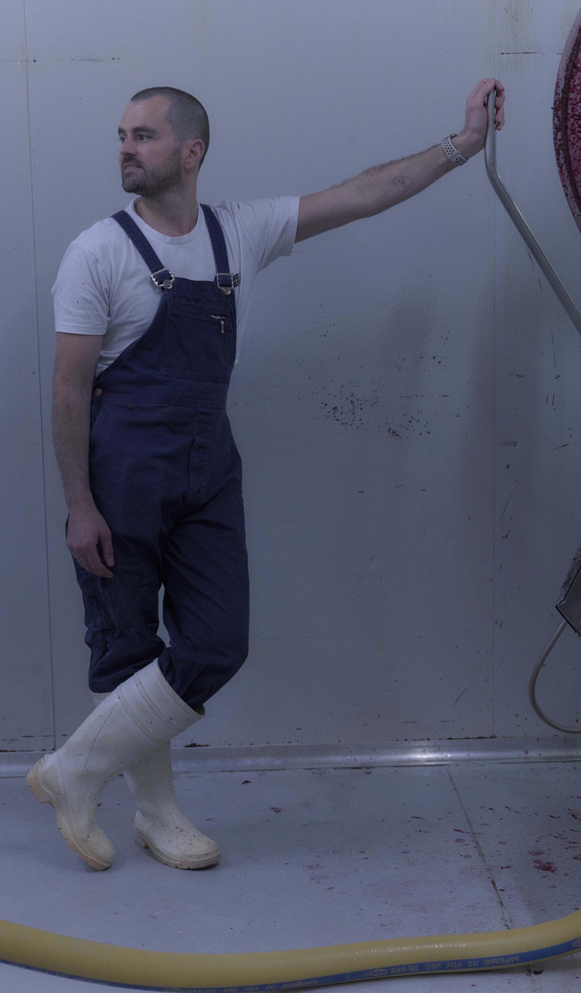 A man dressed in blue overalls, a white t-shirt, and white rubber boots stands in a sterile, industrial room next to a large piece of stainless steel equipment. He leans casually against the machine, which has traces of red liquid, possibly from food processing. The scene is minimalist, with the white walls and floor emphasising the industrial nature of the environment.