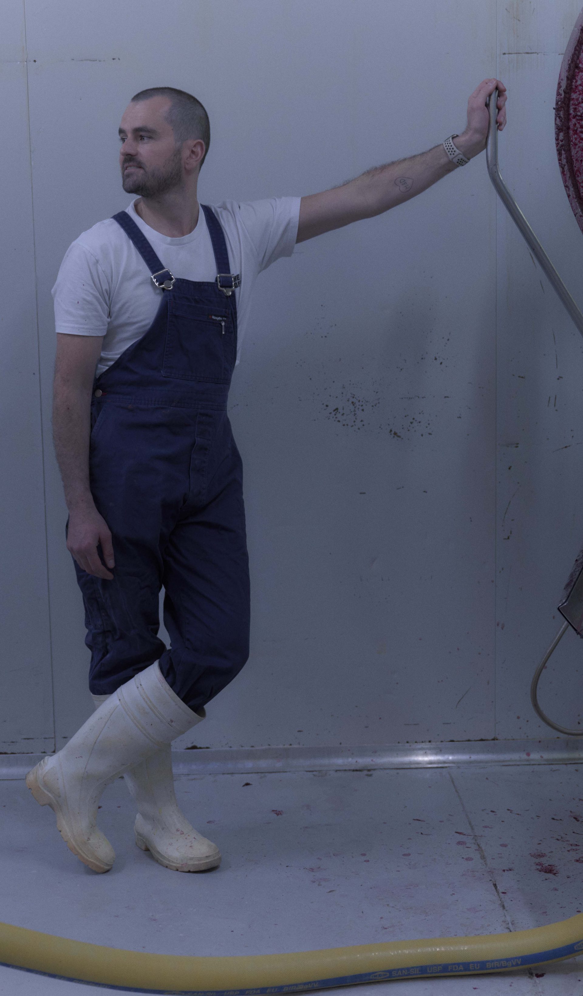 A man dressed in blue overalls, a white t-shirt, and white rubber boots stands in a sterile, industrial room next to a large piece of stainless steel equipment. He leans casually against the machine, which has traces of red liquid, possibly from food processing. The scene is minimalist, with the white walls and floor emphasising the industrial nature of the environment.