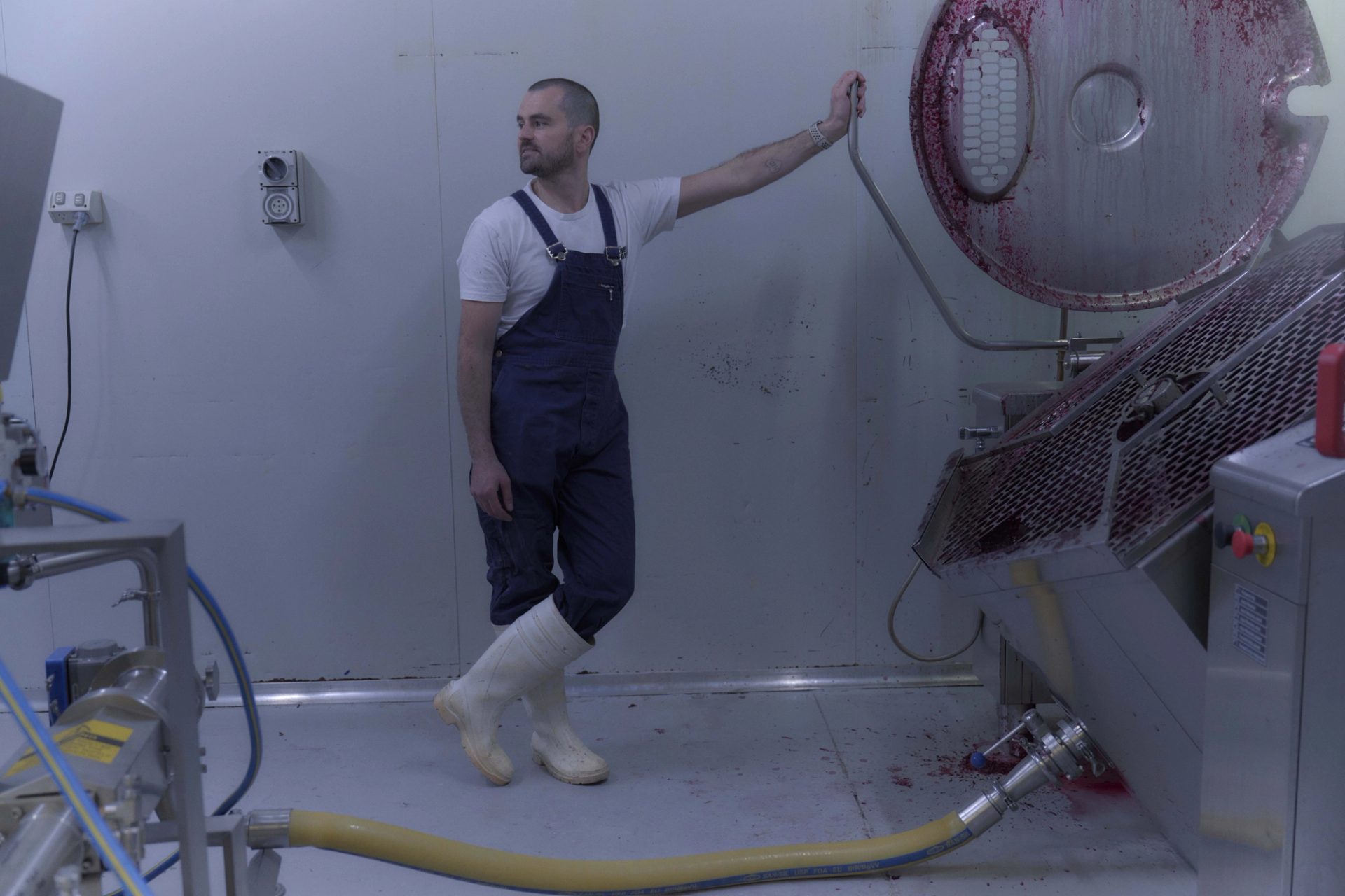 A man dressed in blue overalls, a white t-shirt, and white rubber boots stands in a sterile, industrial room next to a large piece of stainless steel equipment. He leans casually against the machine, which has traces of red liquid, possibly from food processing. The scene is minimalist, with the white walls and floor emphasising the industrial nature of the environment.