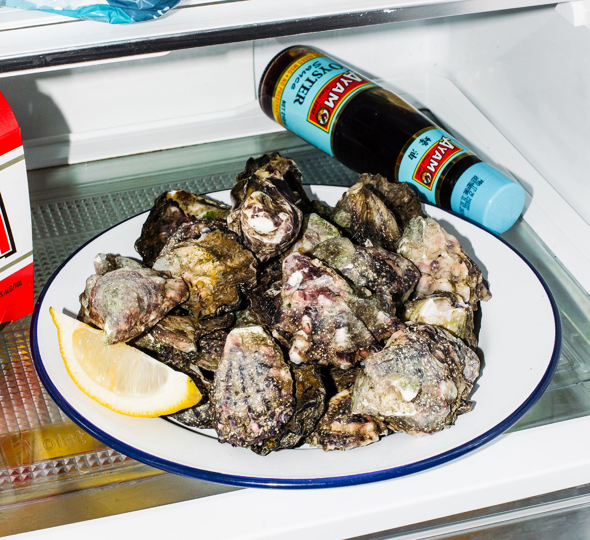 A plate of unchecked oysters with a slice of lemon on a white plate in a fridge.