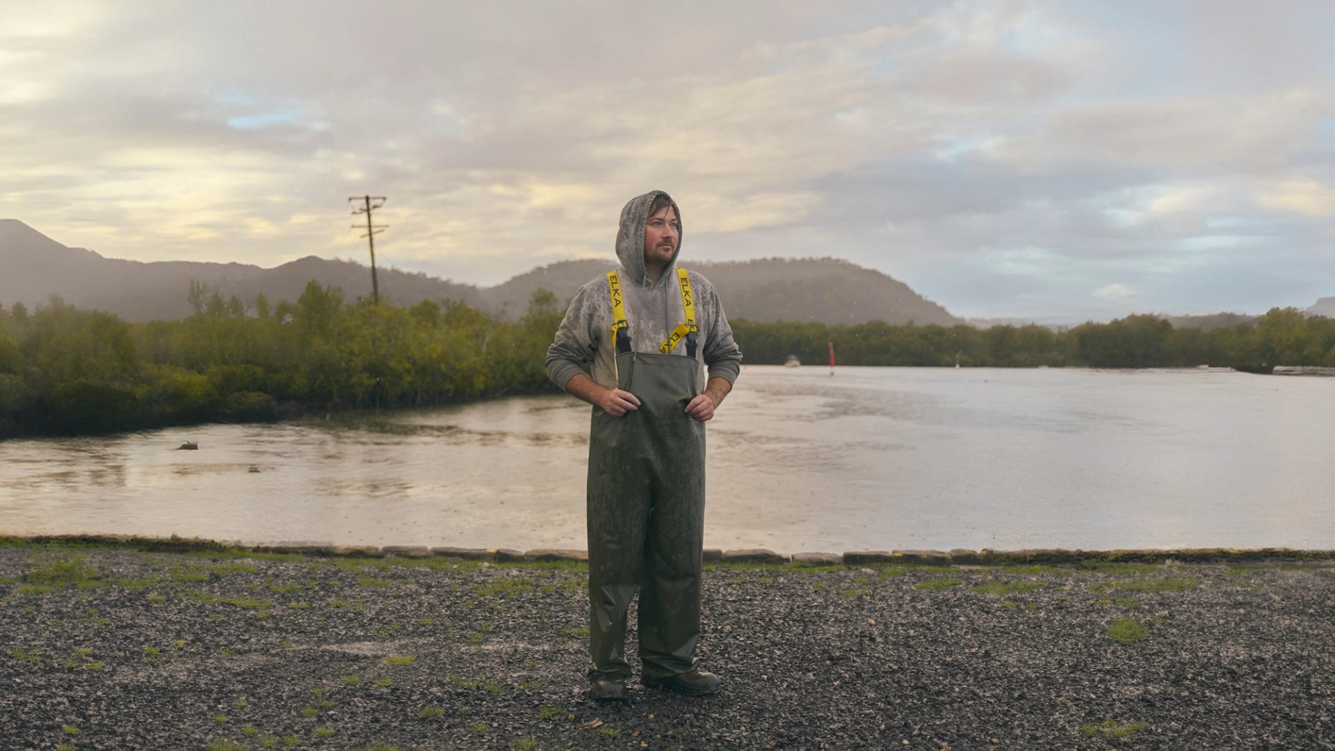 A man stands outdoors near a calm body of water, dressed in heavy-duty rain gear with a hooded sweatshirt underneath. The weather is overcast, with light rain falling, and the landscape behind him includes green hills and a misty sky. The scene has a quiet, reflective atmosphere, with the man appearing thoughtful as he looks into the distance.