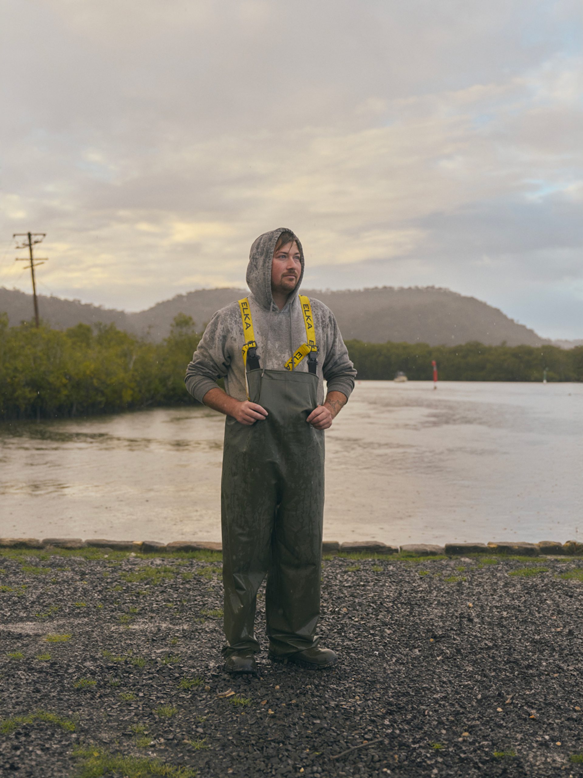 A man stands outdoors near a calm body of water, dressed in heavy-duty rain gear with a hooded sweatshirt underneath. The weather is overcast, with light rain falling, and the landscape behind him includes green hills and a misty sky. The scene has a quiet, reflective atmosphere, with the man appearing thoughtful as he looks into the distance.