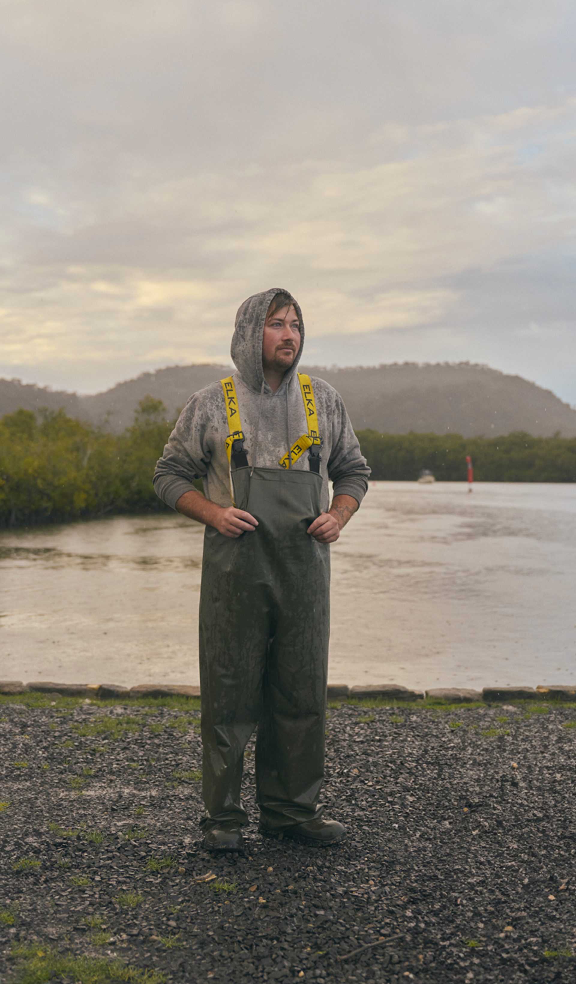 A man stands outdoors near a calm body of water, dressed in heavy-duty rain gear with a hooded sweatshirt underneath. The weather is overcast, with light rain falling, and the landscape behind him includes green hills and a misty sky. The scene has a quiet, reflective atmosphere, with the man appearing thoughtful as he looks into the distance.