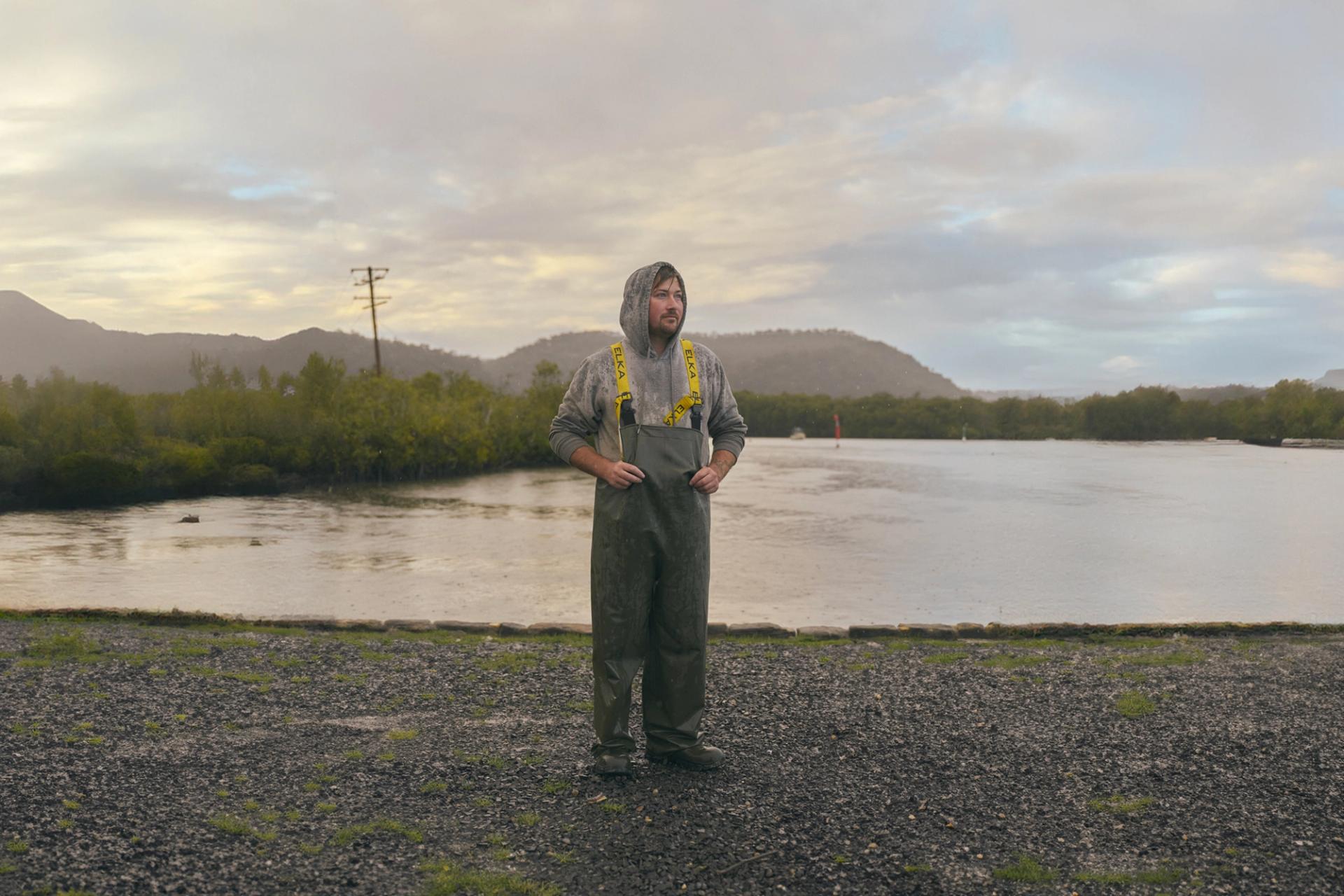 A man stands outdoors near a calm body of water, dressed in heavy-duty rain gear with a hooded sweatshirt underneath. The weather is overcast, with light rain falling, and the landscape behind him includes green hills and a misty sky. The scene has a quiet, reflective atmosphere, with the man appearing thoughtful as he looks into the distance.