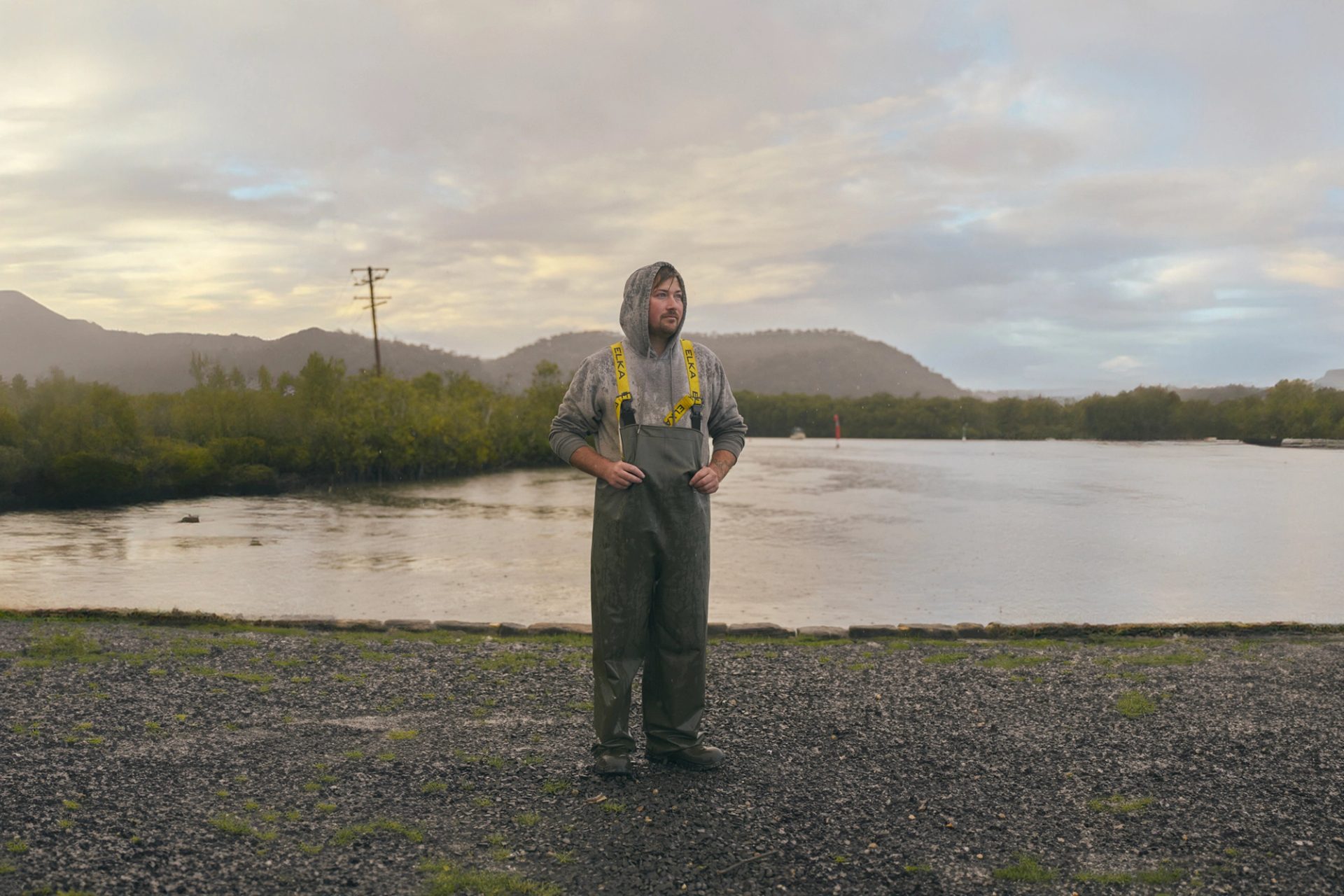 A man stands outdoors near a calm body of water, dressed in heavy-duty rain gear with a hooded sweatshirt underneath. The weather is overcast, with light rain falling, and the landscape behind him includes green hills and a misty sky. The scene has a quiet, reflective atmosphere, with the man appearing thoughtful as he looks into the distance.