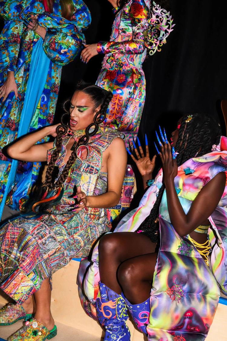Models wearing multicoloured outfits pose for a photo sitting down on wooden benches