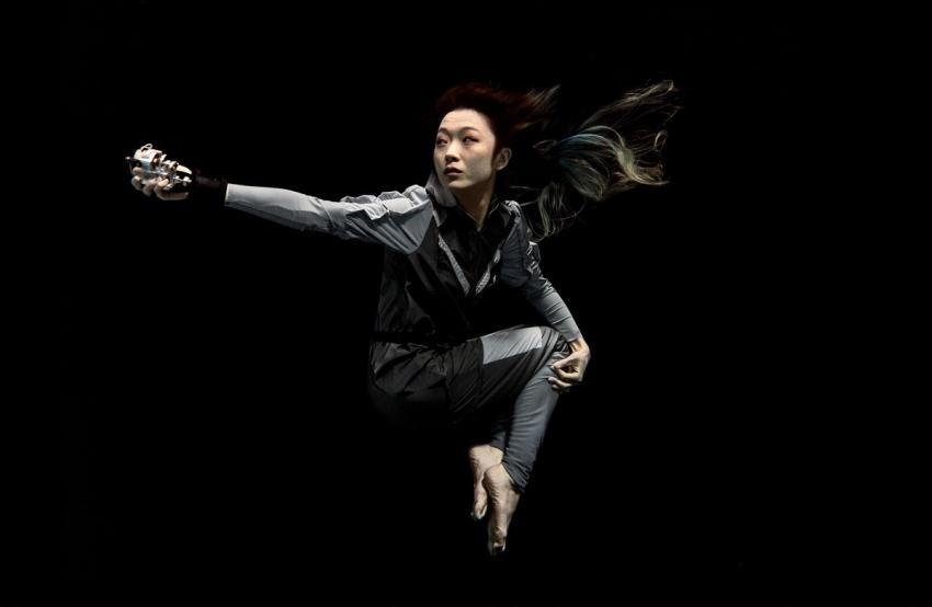 Artist Xin Liu floating with arm outstretched against a black background. She wears a full-length grey body suit with long sleeves with bare feet and hands.