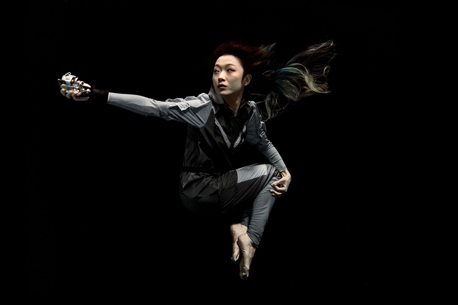 Artist Xin Liu floating with arm outstretched against a black background. She wears a full-length grey body suit with long sleeves with bare feet and hands.