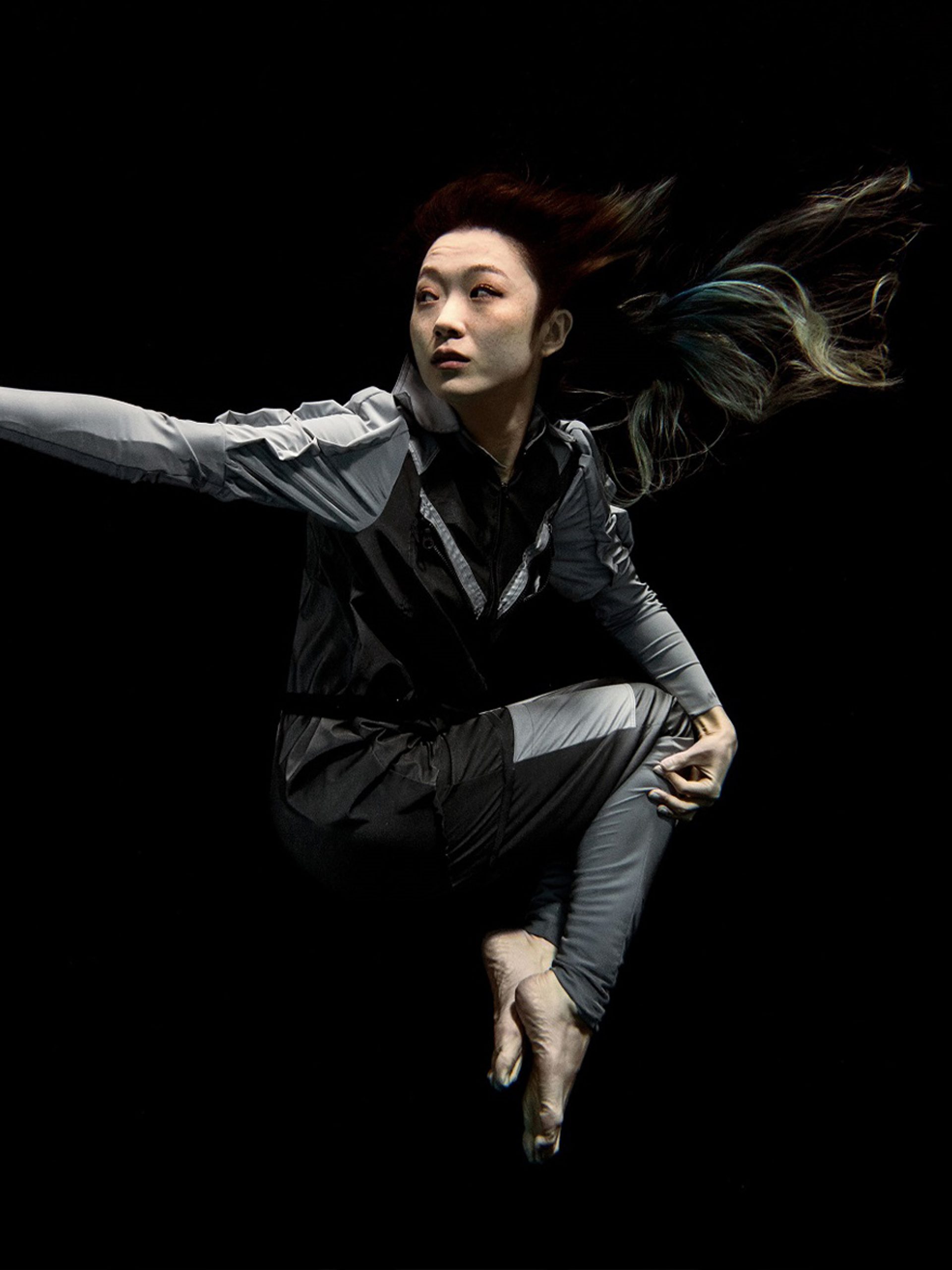 Artist Xin Liu floating with arm outstretched against a black background. She wears a full-length grey body suit with long sleeves with bare feet and hands.