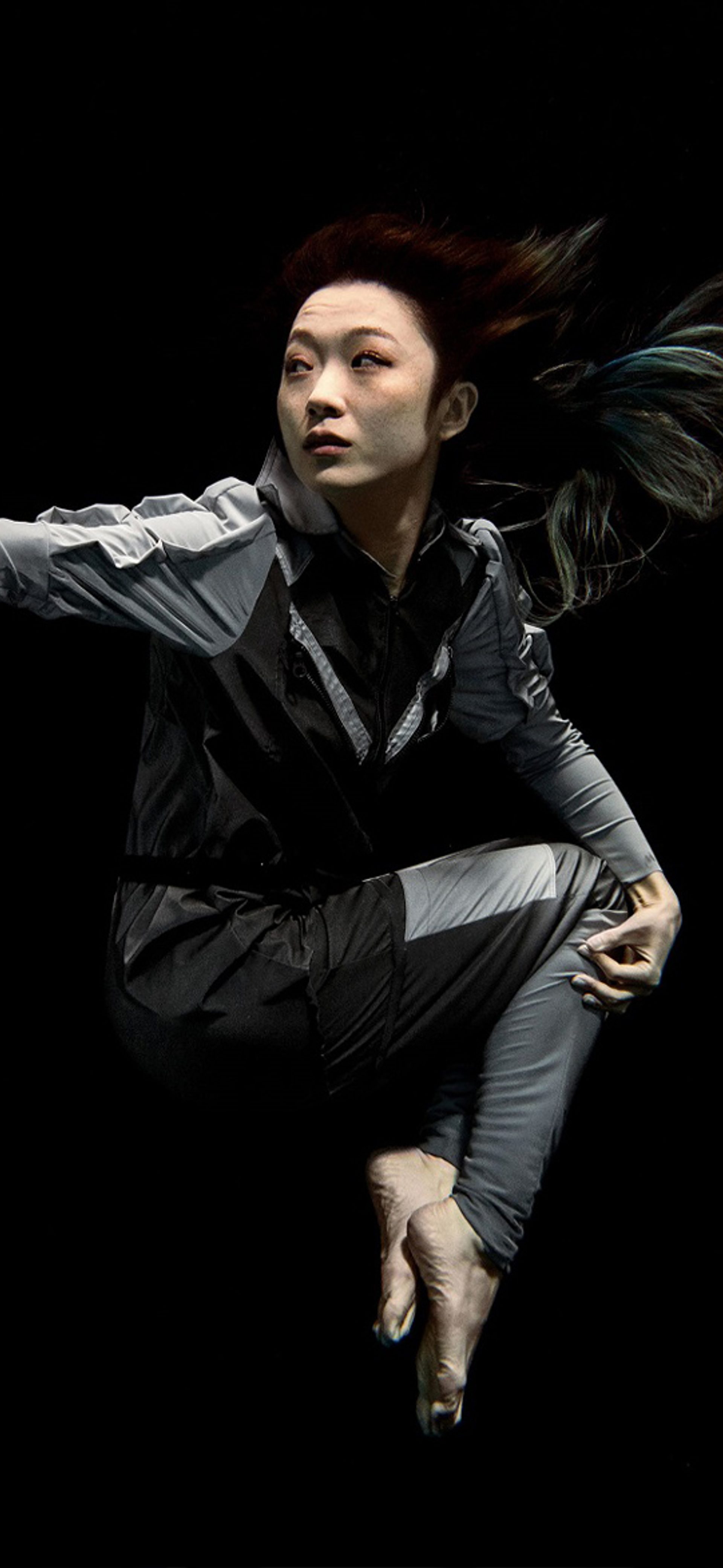 Artist Xin Liu floating with arm outstretched against a black background. She wears a full-length grey body suit with long sleeves with bare feet and hands.