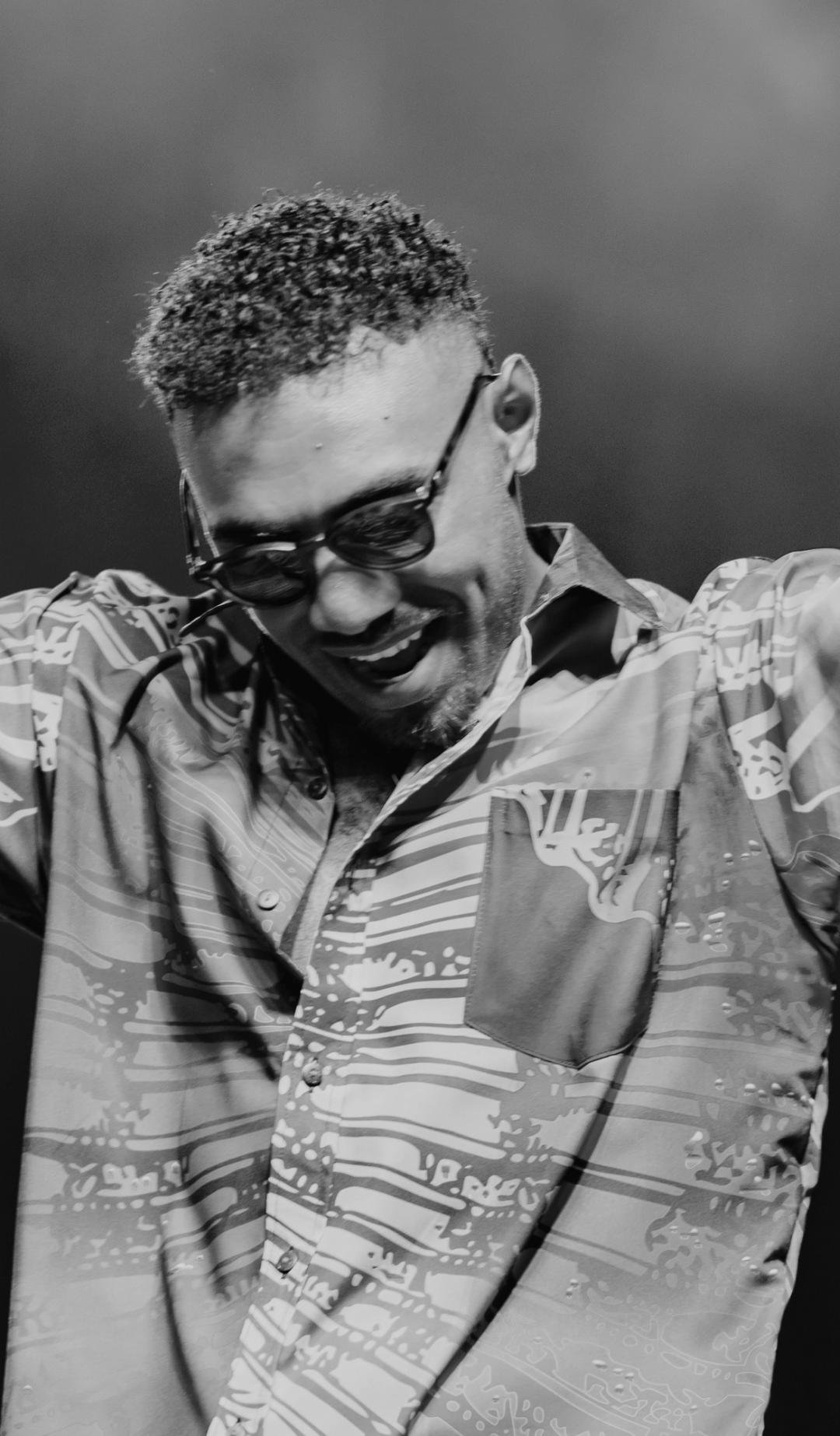 Black and white photo of Jay Tauleka performing on stage wearing sunglasses with his hands up