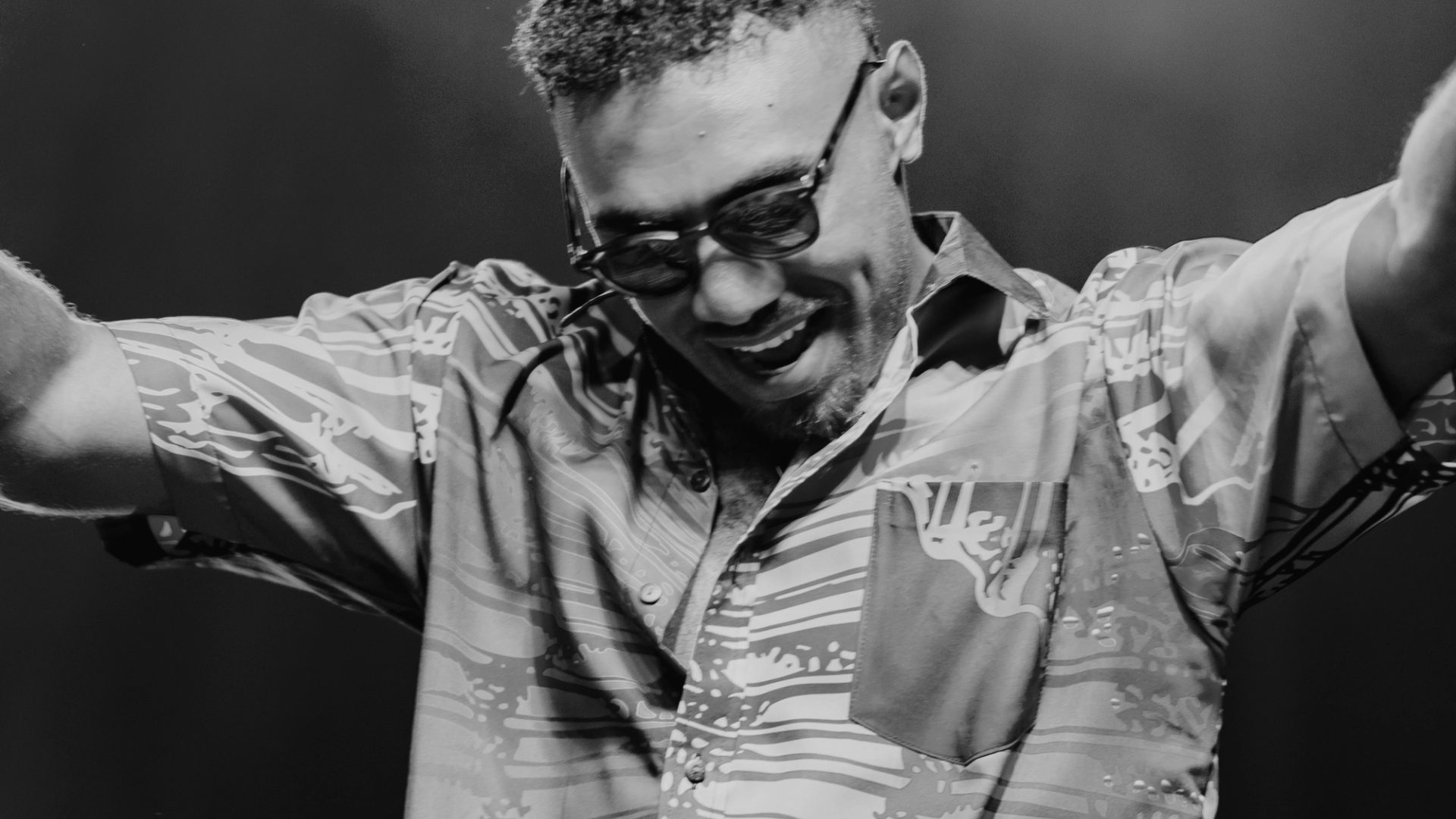Black and white photo of Jay Tauleka performing on stage wearing sunglasses with his hands up