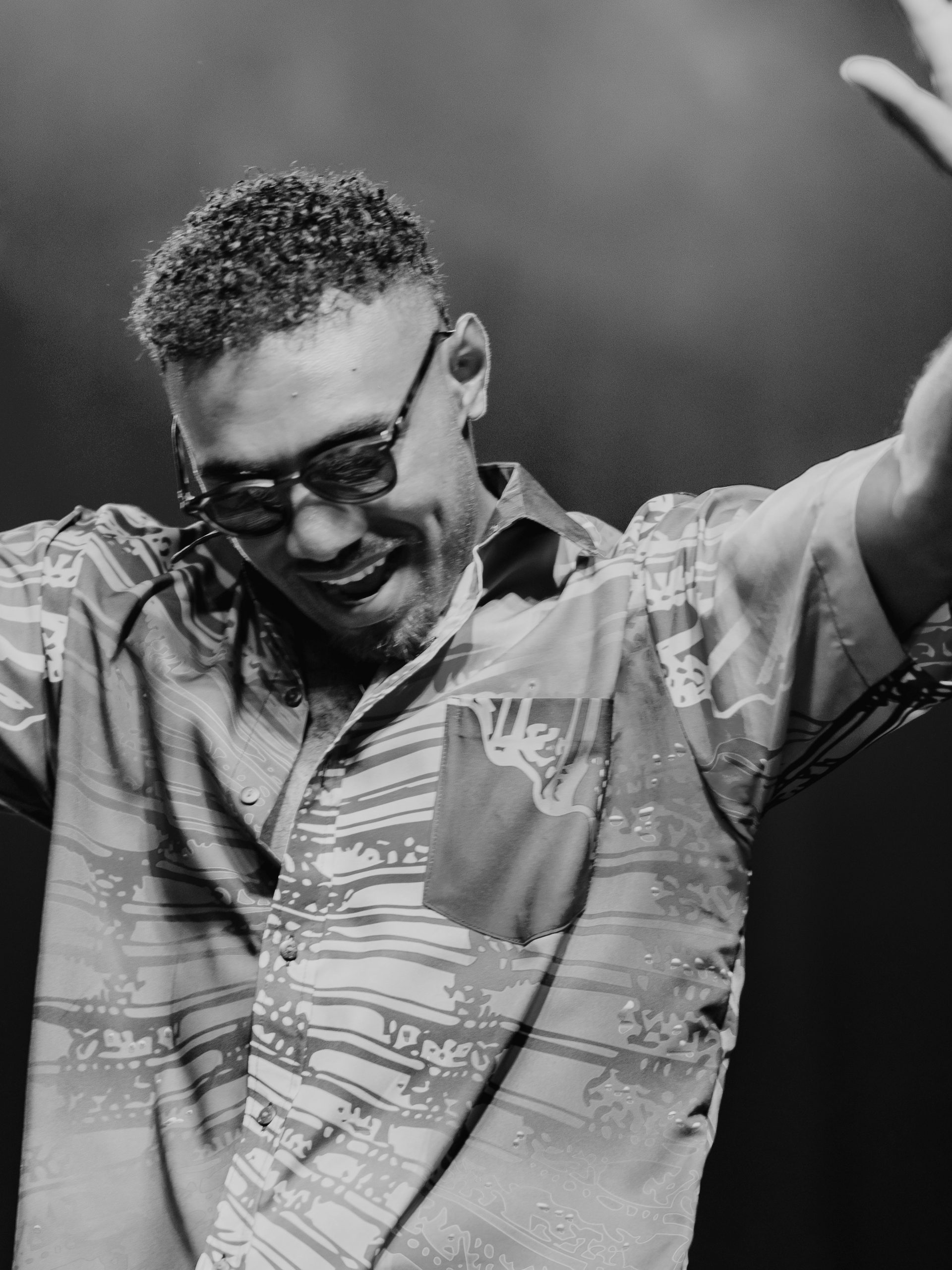 Black and white photo of Jay Tauleka performing on stage wearing sunglasses with his hands up