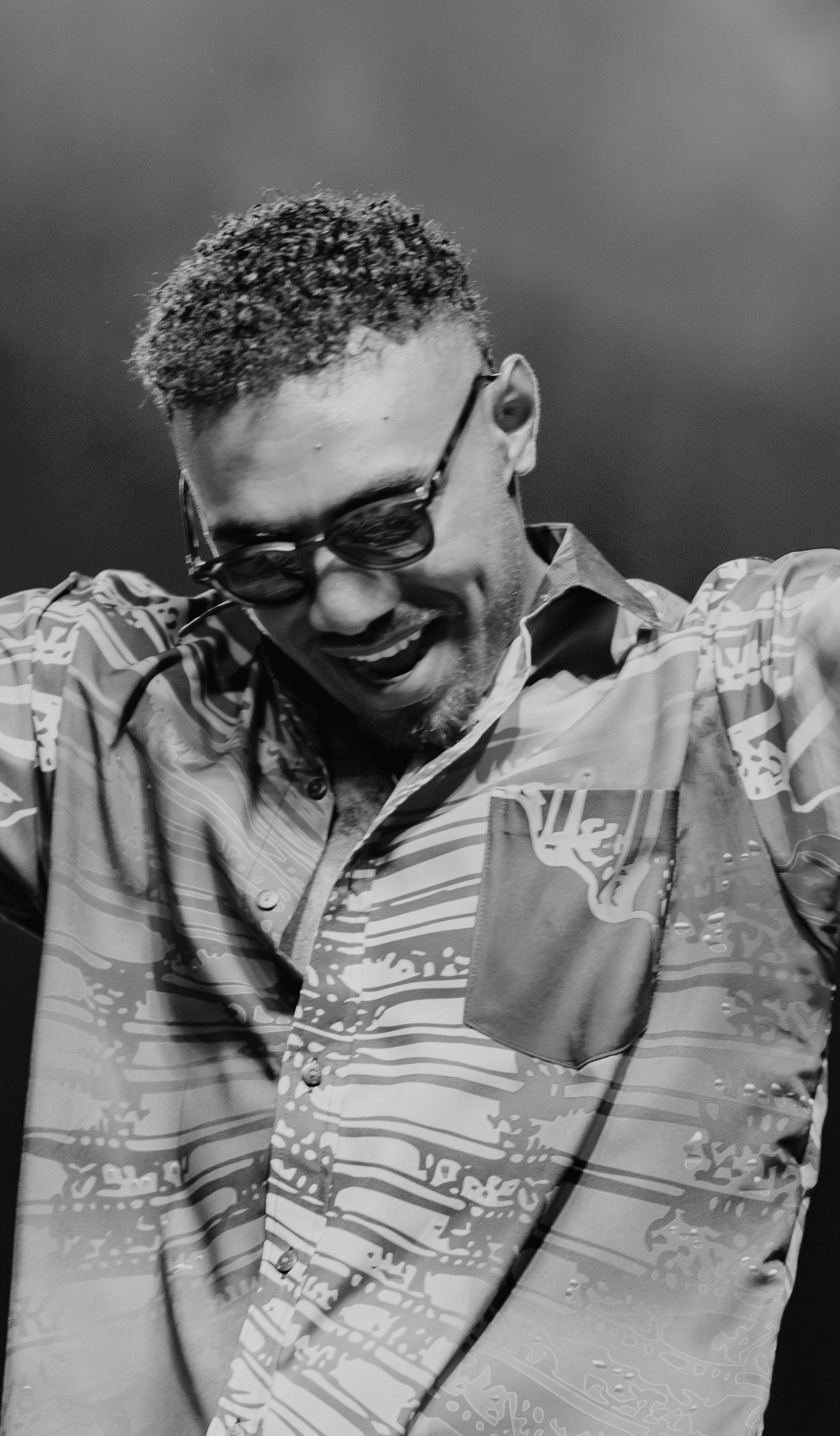 Black and white photo of Jay Tauleka performing on stage wearing sunglasses with his hands up