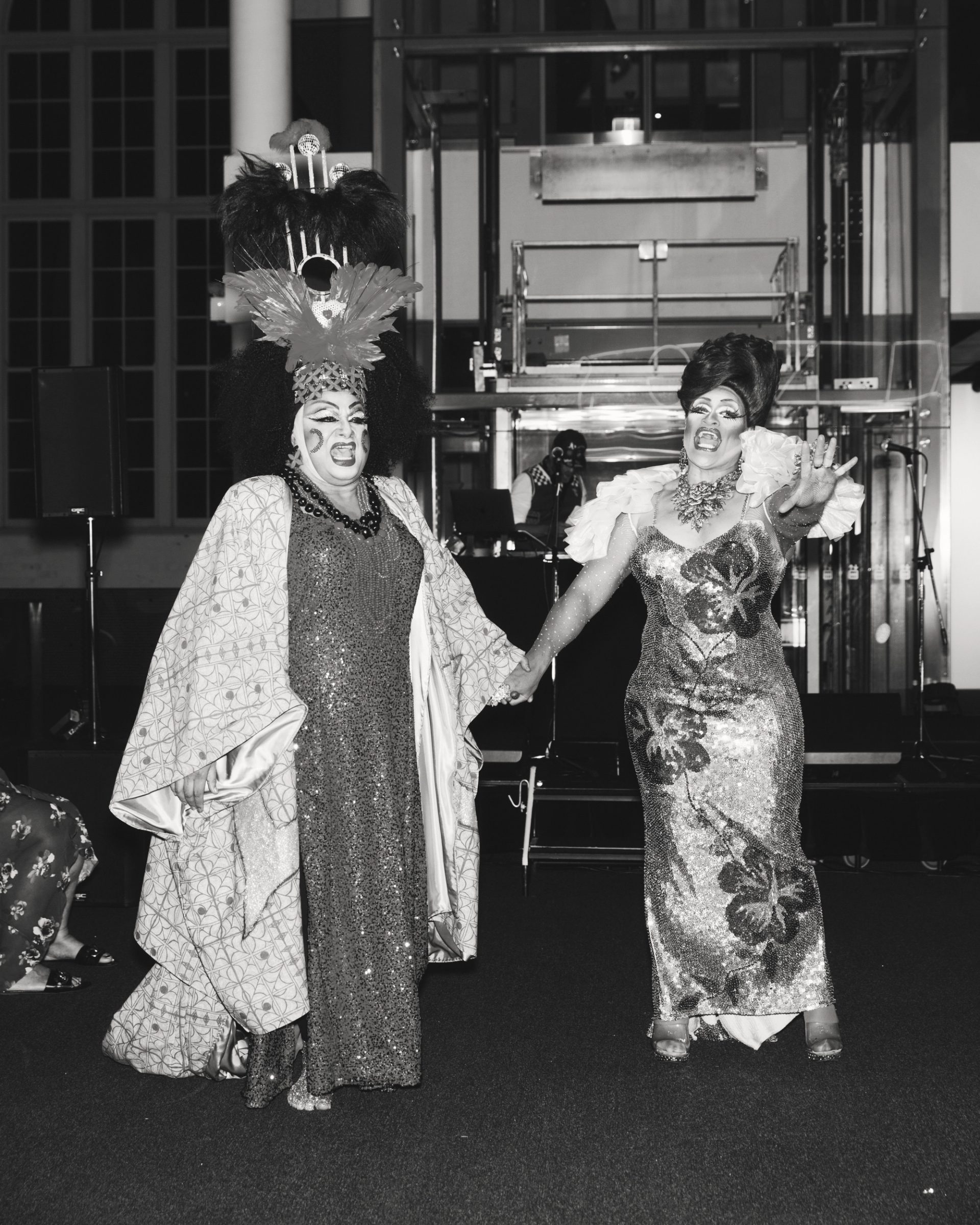 Two drag queens standing close together mid performance.