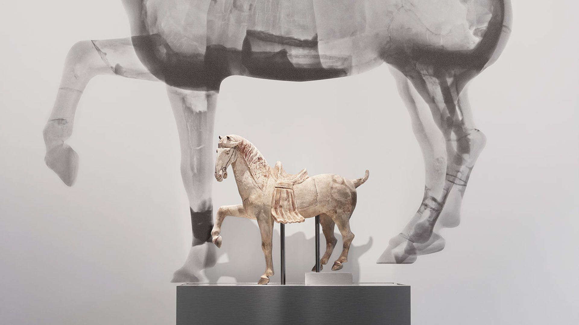 Small horse figure on a plinth in front of a large black and white neutron tomography scan of the same horse figure.