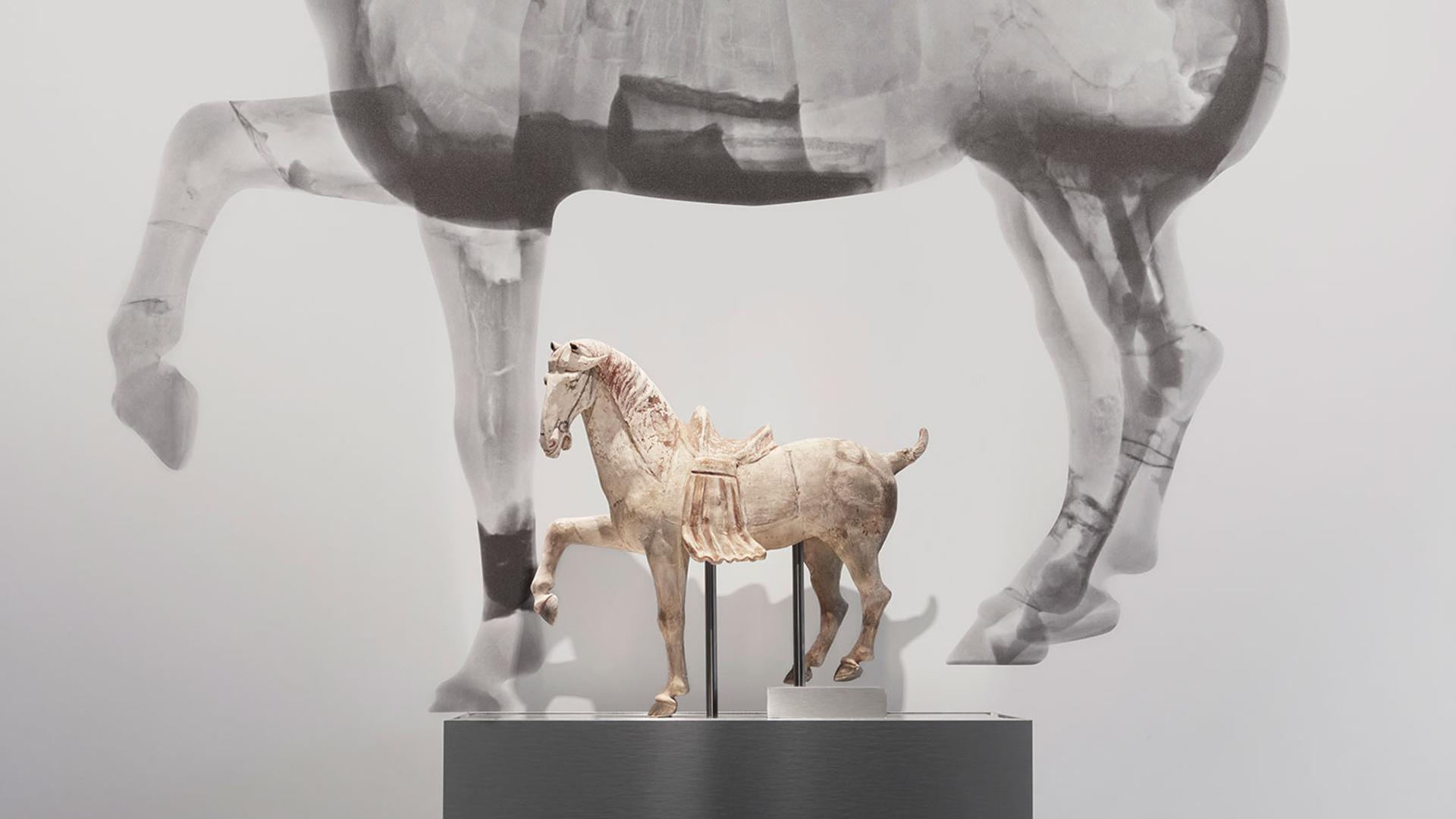 Small horse figure on a plinth in front of a large black and white neutron tomography scan of the same horse figure.
