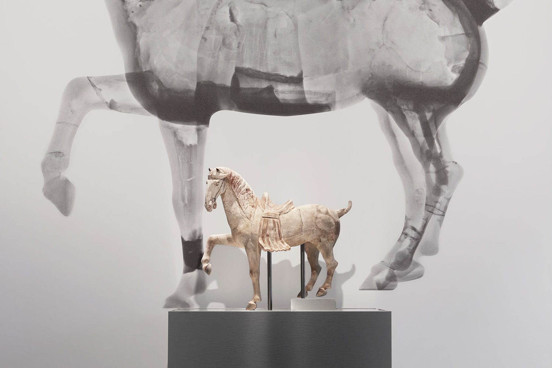 Small horse figure on a plinth in front of a large black and white neutron tomography scan of the same horse figure.