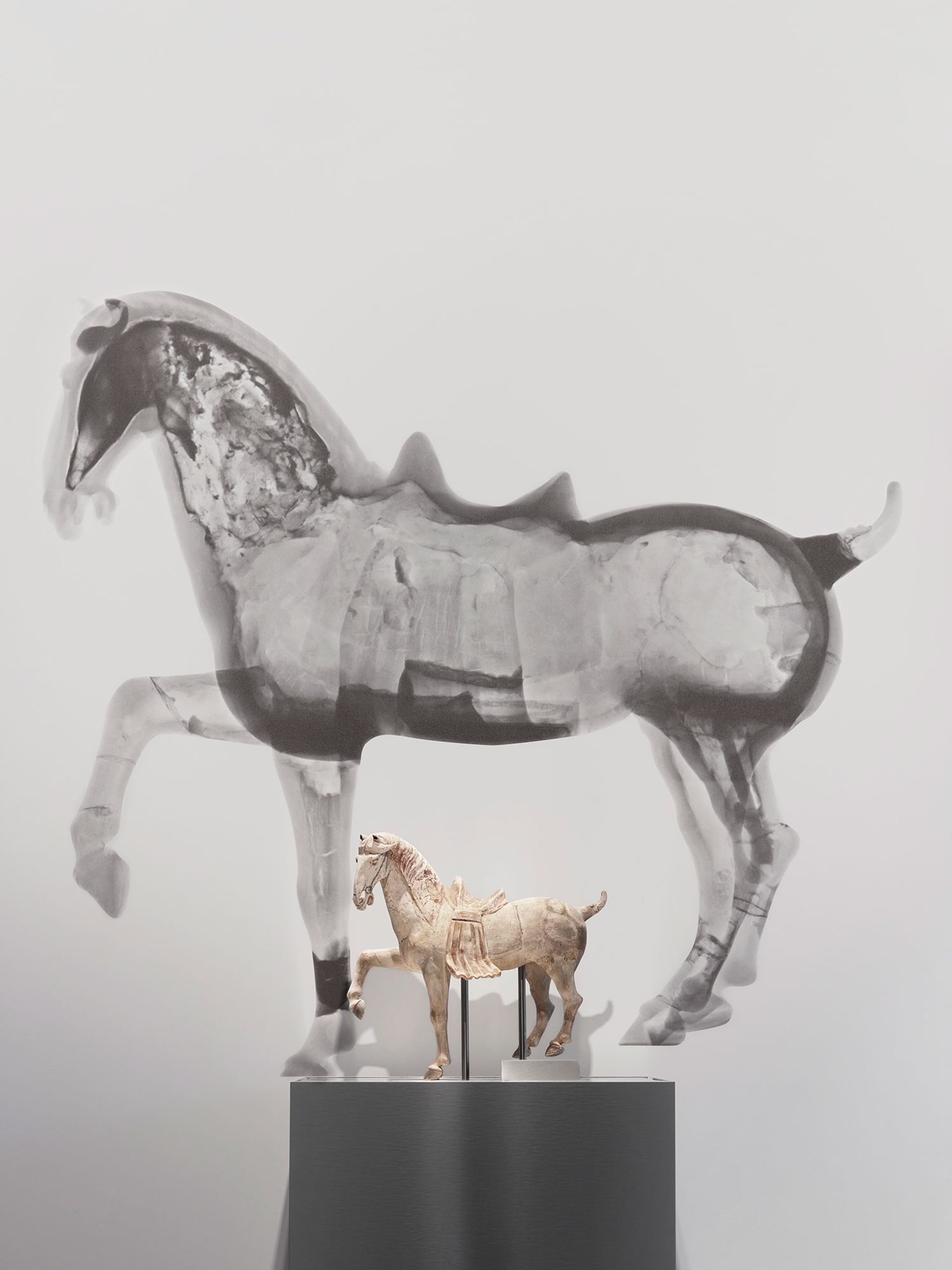 Small horse figure on a plinth in front of a large black and white neutron tomography scan of the same horse figure.