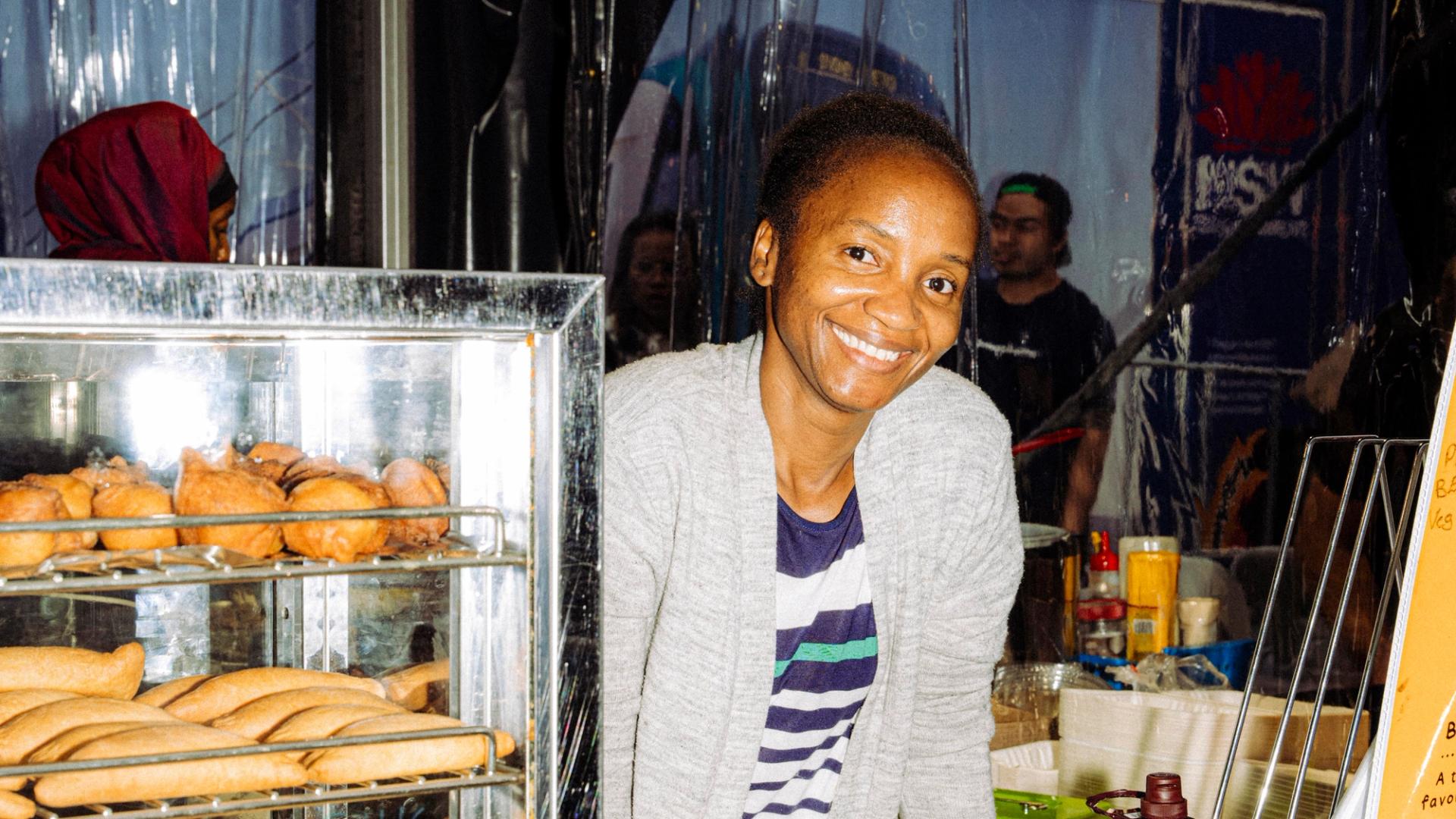 A smiling person stands behind a food stall displaying trays of baked goods in a glass warmer. Drink bottles and condiments are on the counter in front of them.