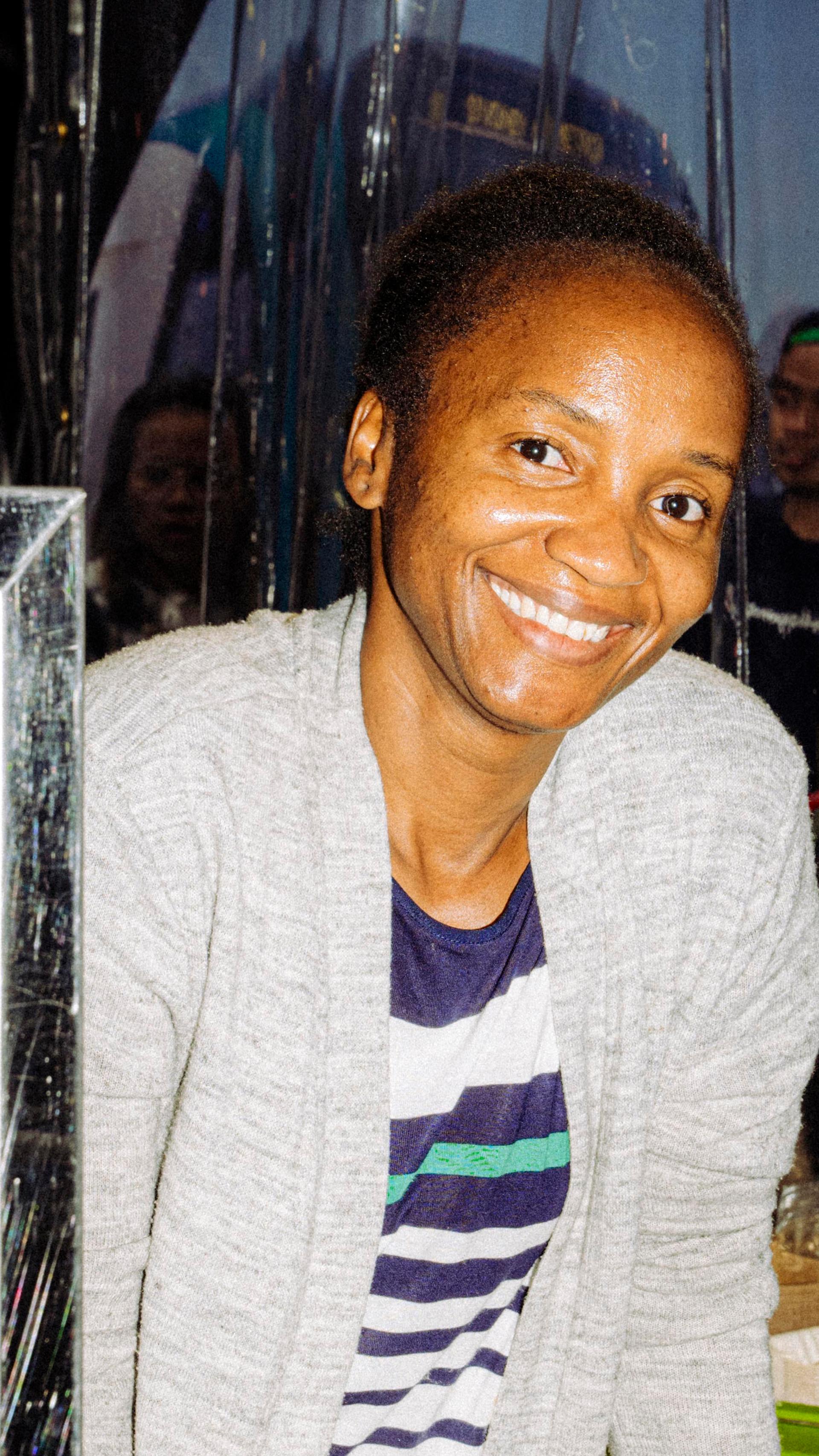 A smiling person stands behind a food stall displaying trays of baked goods in a glass warmer. Drink bottles and condiments are on the counter in front of them.