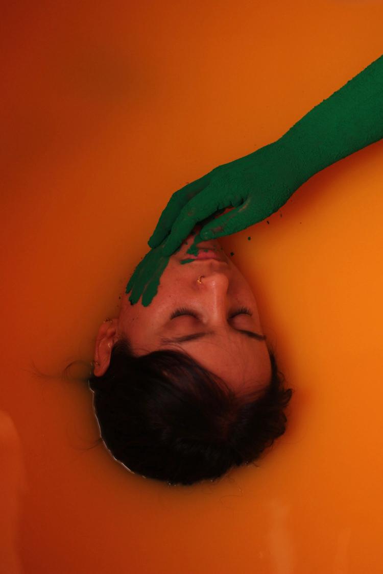 Face in orange liquid bath, with green arm and hand reaching for face from top right of frame.
