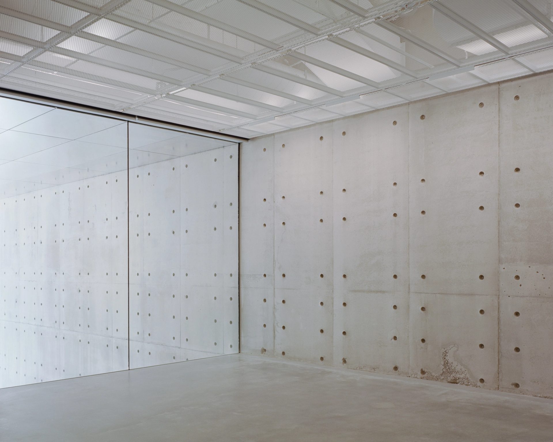 A large room with a concrete floor, the wall on the left is glass and the back wall is patterned concrete.