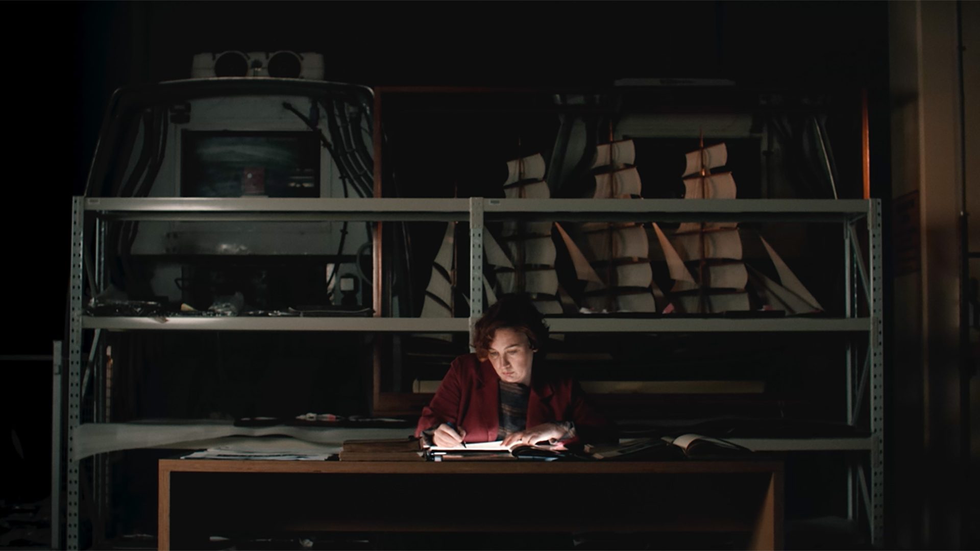 Figure sits at a desk writing on an illuminated surface, shelves are in the background.