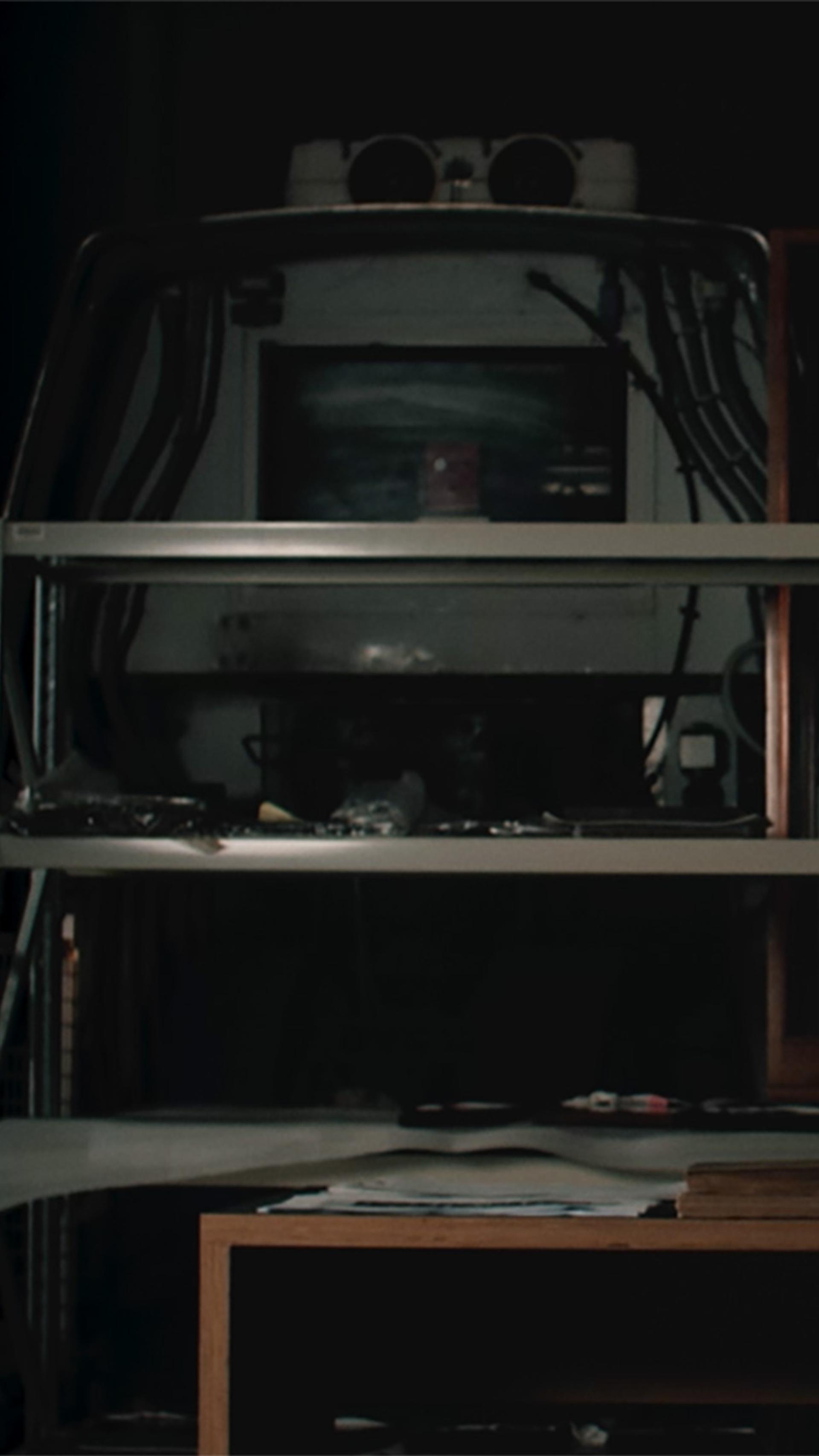 Figure sits at a desk writing on an illuminated surface, shelves are in the background.