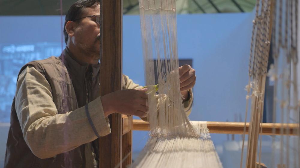 A figure works a wooden loom strung with beige threads, they cut the threads with scissors