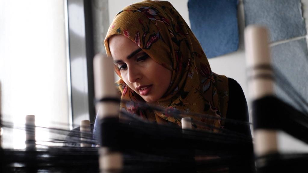 Wajiha Pervez working in her studio. She is looking down at a desk and in the foreground, there are blurred spools of yarn.