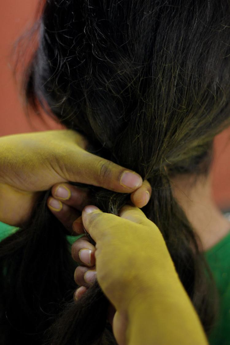 Two hands covered in yellow pigment braid the hair of a person wearing a green top.