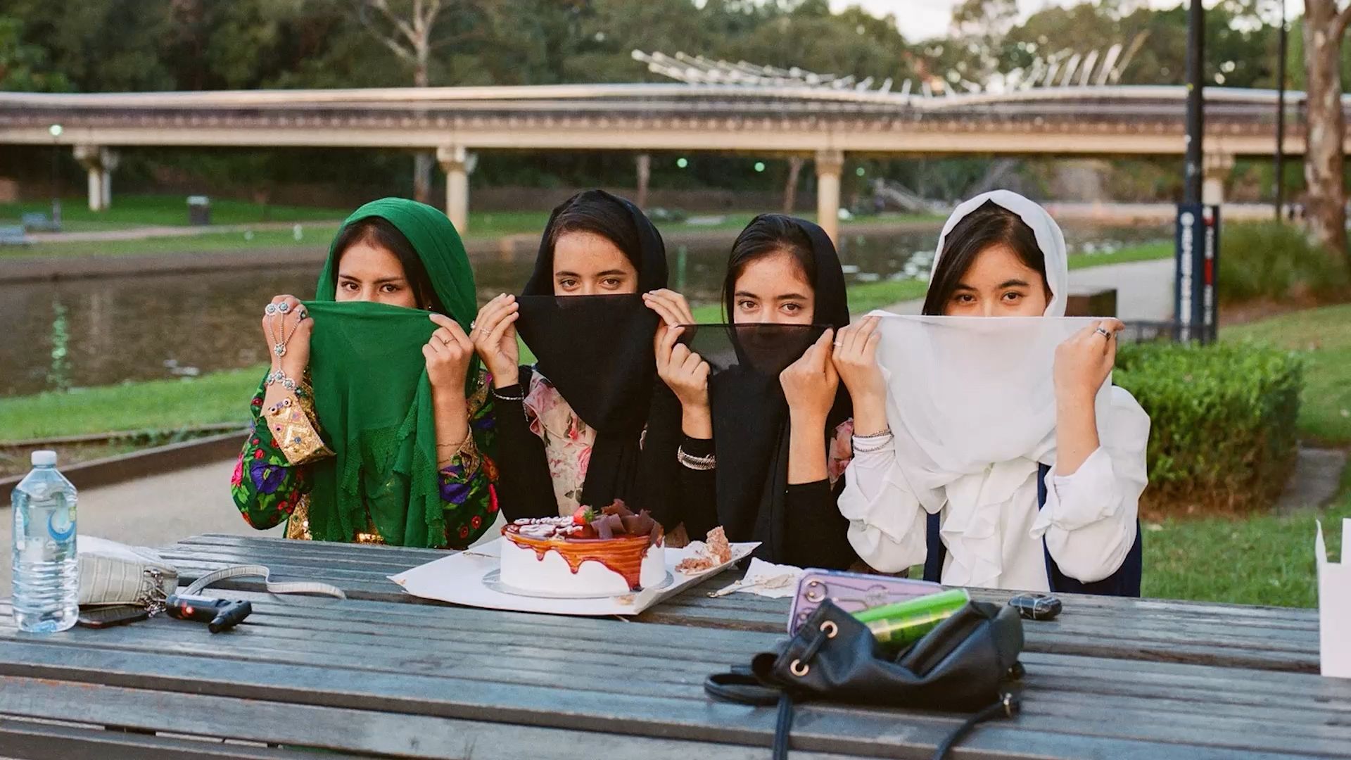 A photograph of four young women sitting outside in the sunshine, at a picnic table, with a delicious looking layer cake in front of them.