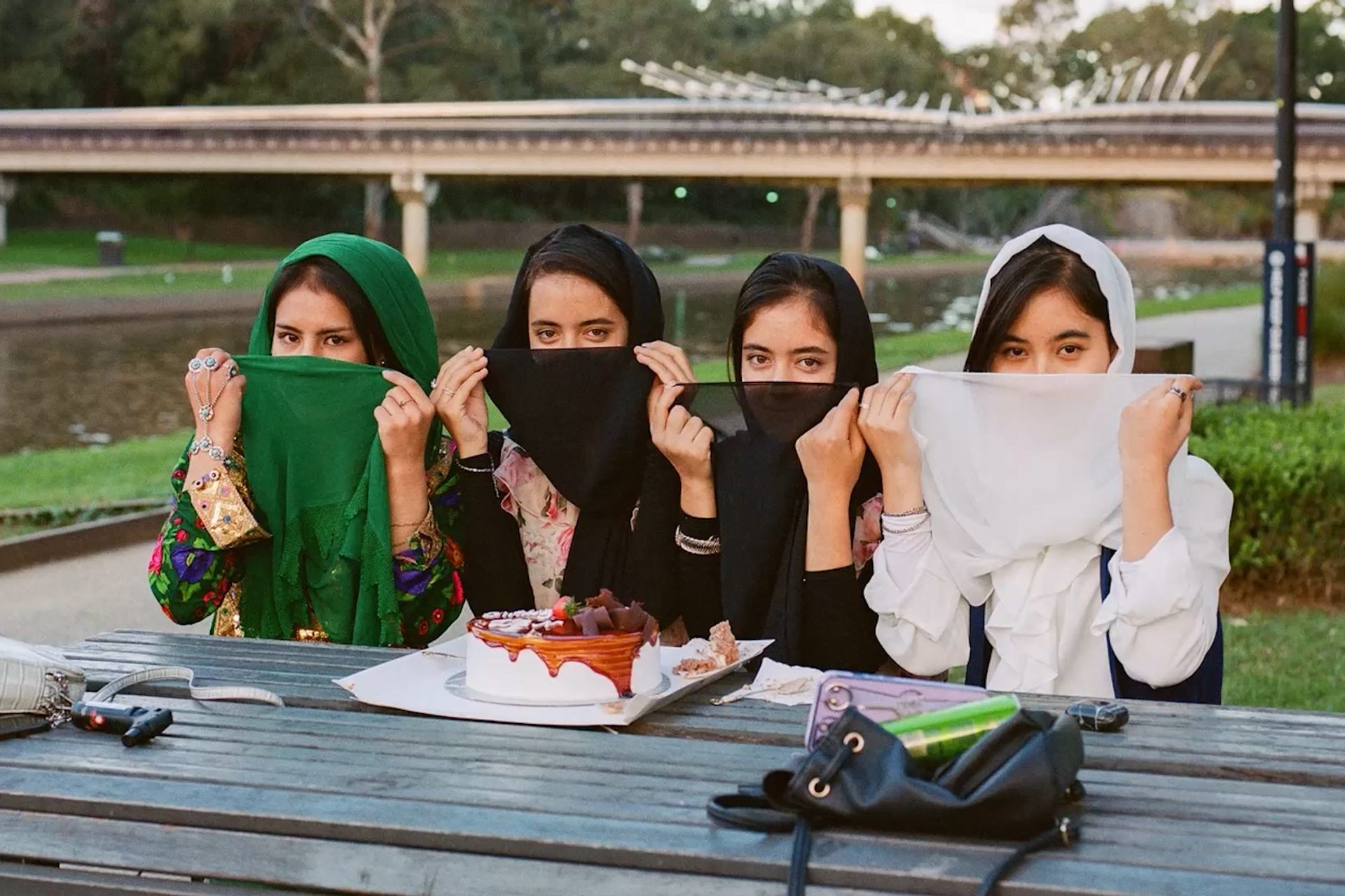 A photograph of four young women sitting outside in the sunshine, at a picnic table, with a delicious looking layer cake in front of them.