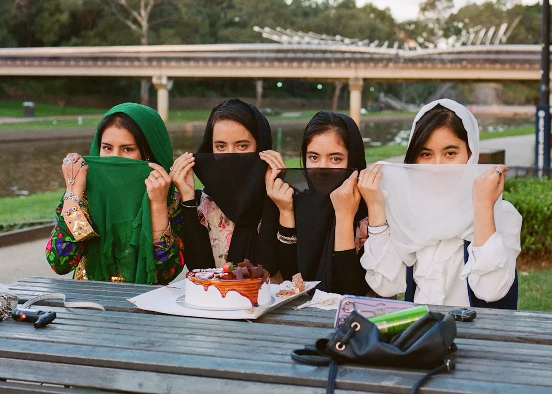 A photograph of four young women sitting outside in the sunshine, at a picnic table, with a delicious looking layer cake in front of them.