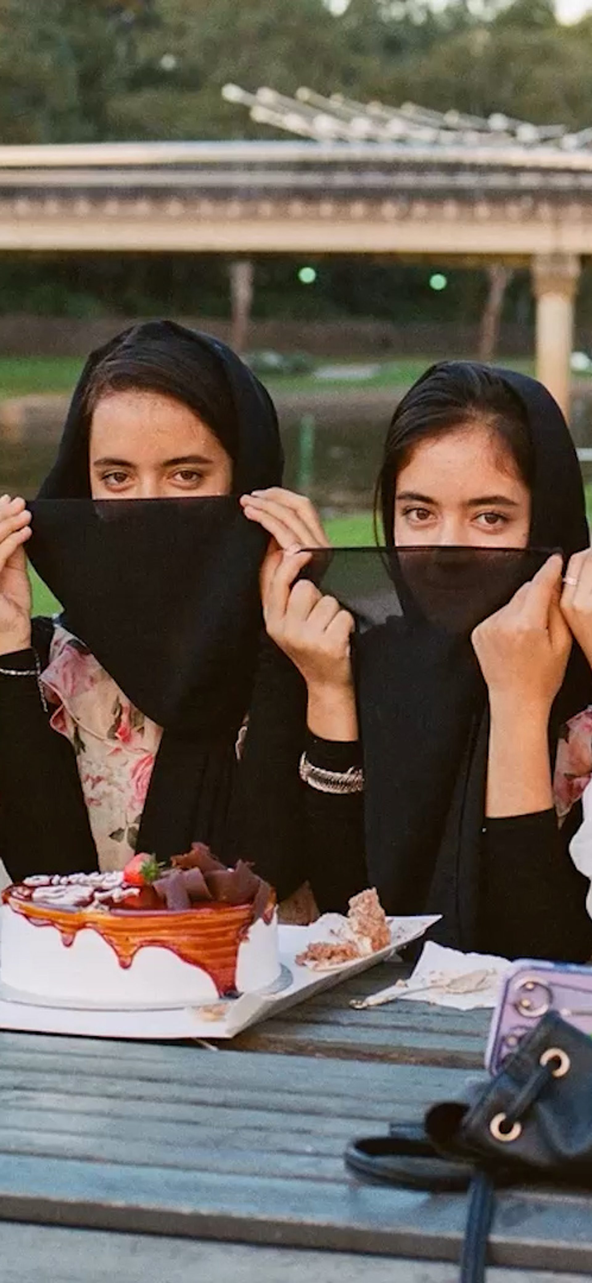 A photograph of four young women sitting outside in the sunshine, at a picnic table, with a delicious looking layer cake in front of them.
