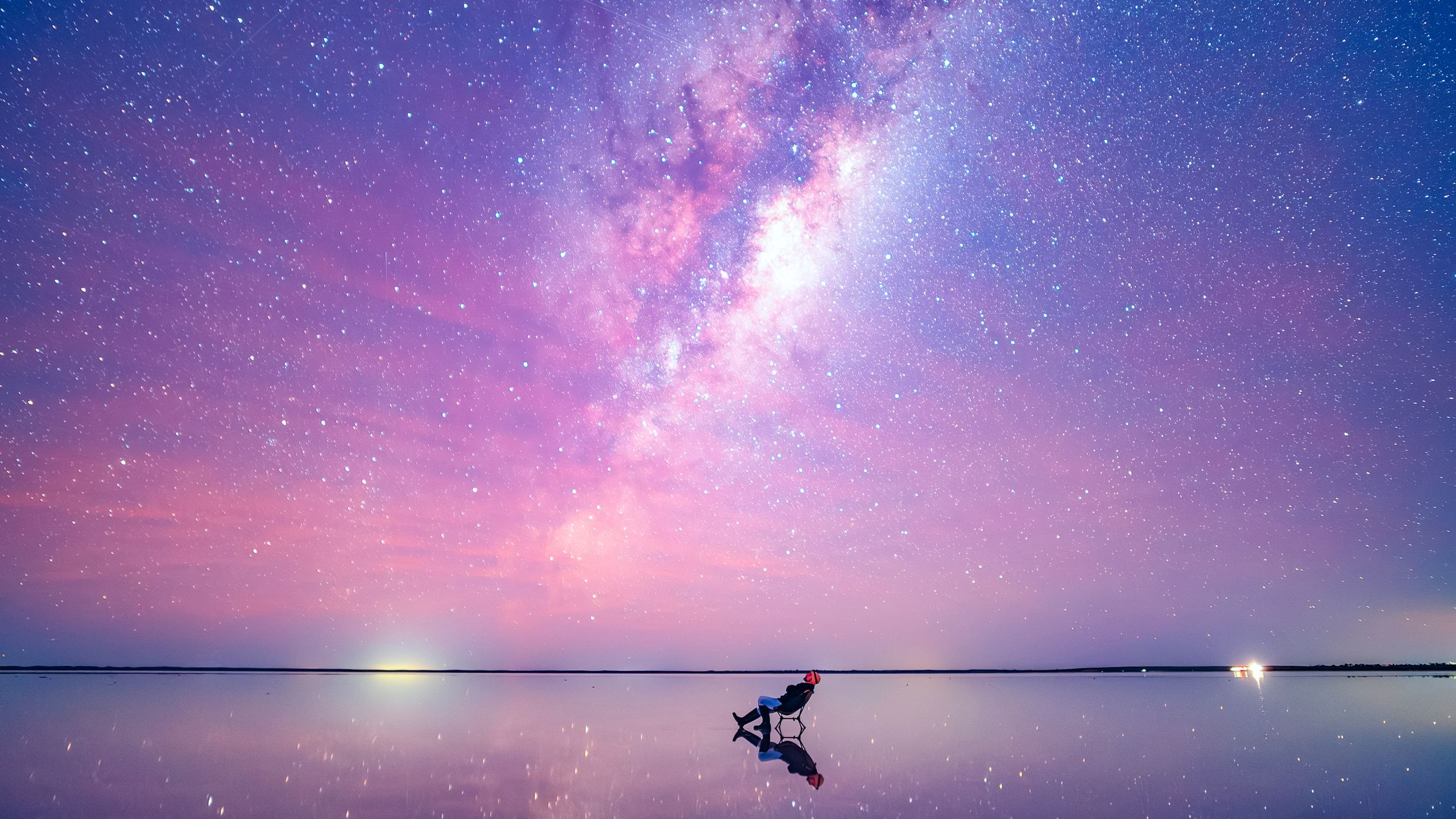 An image of a stargazer, leaning back in a chair, staring up at the sky. The image is shot in an extreme wide: the stargazer looks tiny compared to the bright, colorful and mostly pink sky, and the vast expanse of water that surrounds them. The stars are visible in a long stretch above the stargazer's head.