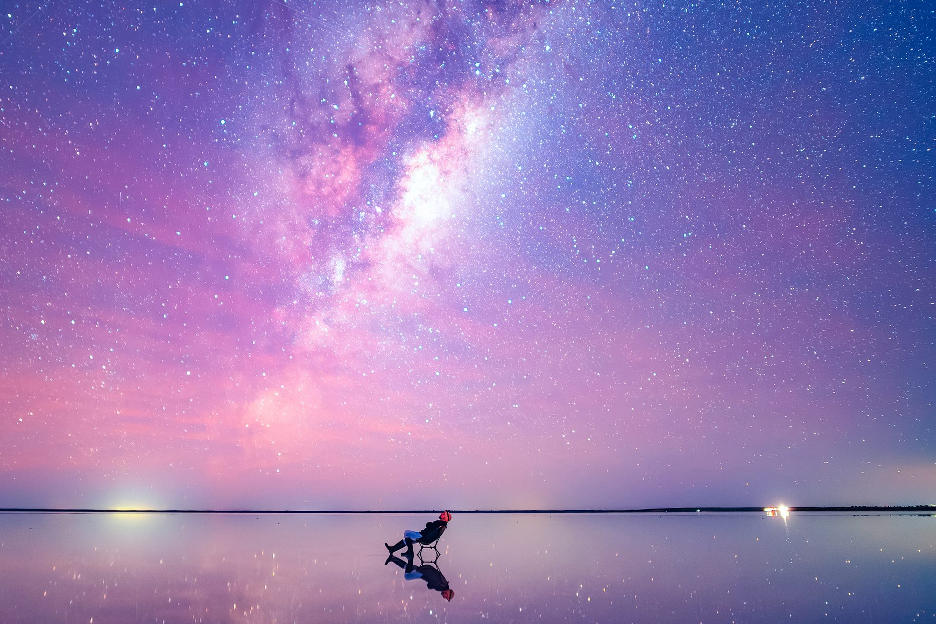 An image of a stargazer, leaning back in a chair, staring up at the sky. The image is shot in an extreme wide: the stargazer looks tiny compared to the bright, colorful and mostly pink sky, and the vast expanse of water that surrounds them. The stars are visible in a long stretch above the stargazer's head.
