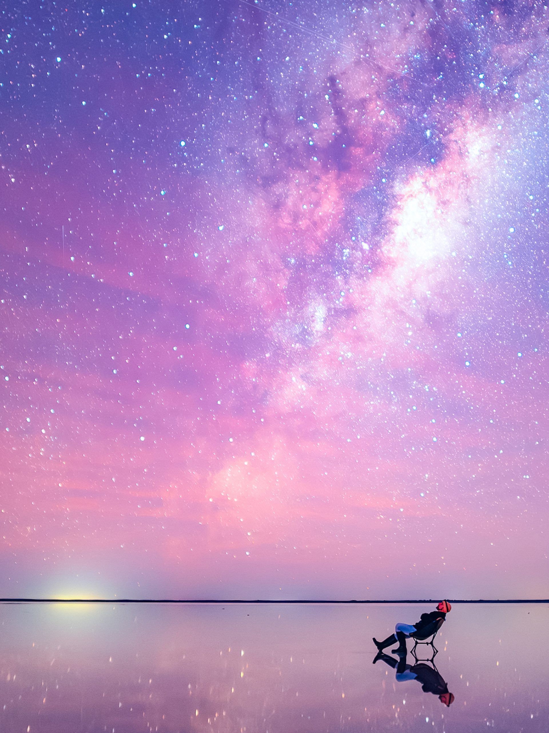 An image of a stargazer, leaning back in a chair, staring up at the sky. The image is shot in an extreme wide: the stargazer looks tiny compared to the bright, colorful and mostly pink sky, and the vast expanse of water that surrounds them. The stars are visible in a long stretch above the stargazer's head.