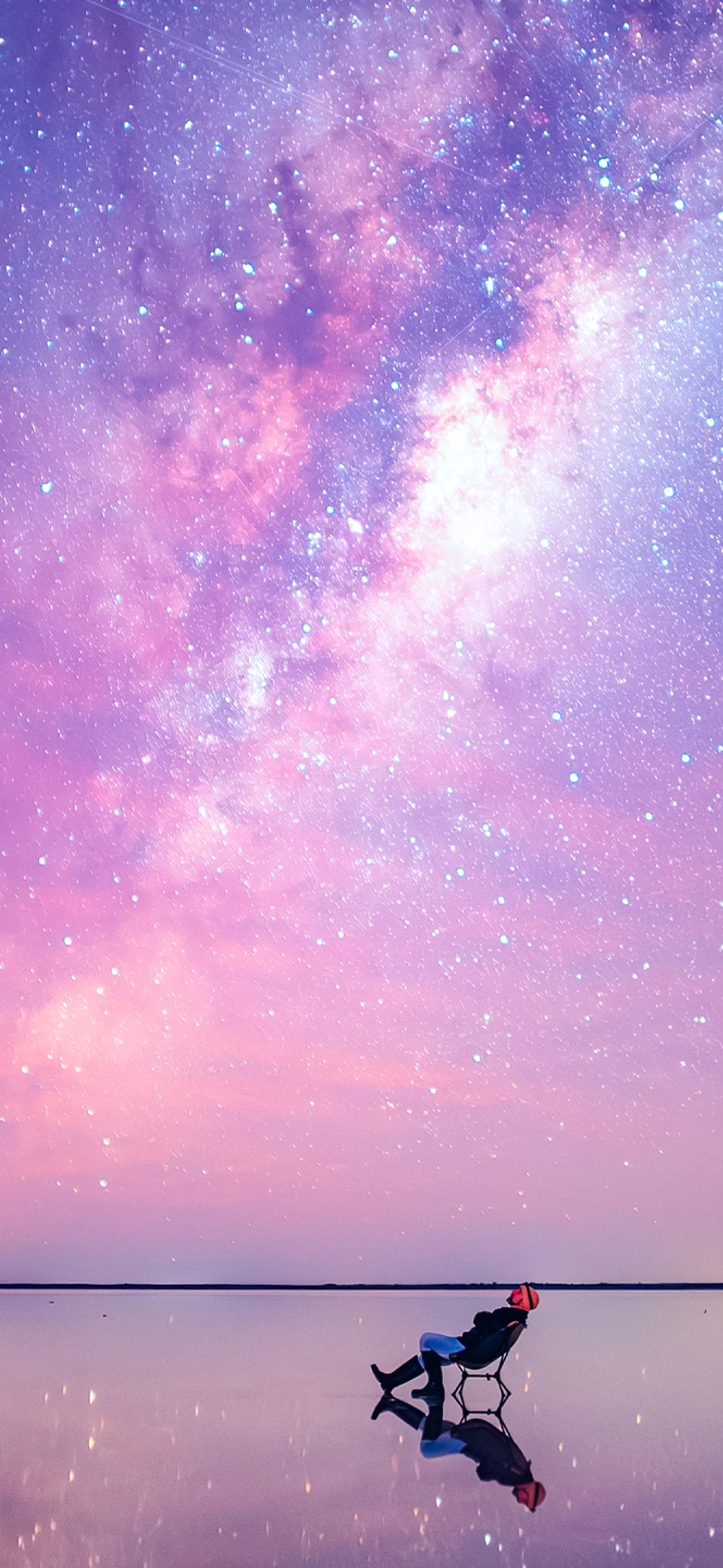 An image of a stargazer, leaning back in a chair, staring up at the sky. The image is shot in an extreme wide: the stargazer looks tiny compared to the bright, colorful and mostly pink sky, and the vast expanse of water that surrounds them. The stars are visible in a long stretch above the stargazer's head.