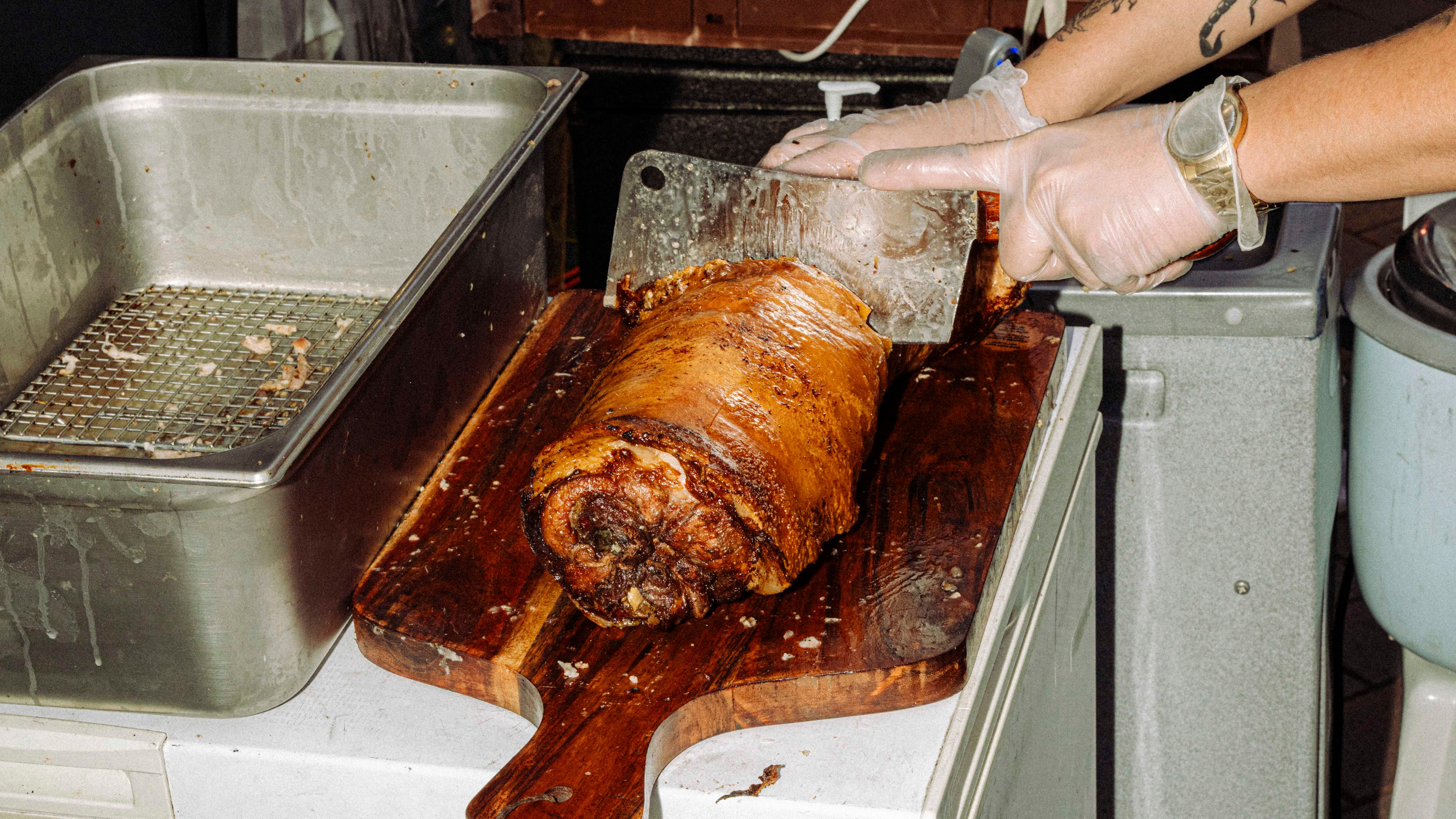 A vendor in gloves slices a large roasted meat piece on a wooden cutting board, with various kitchen tools and containers in the background.