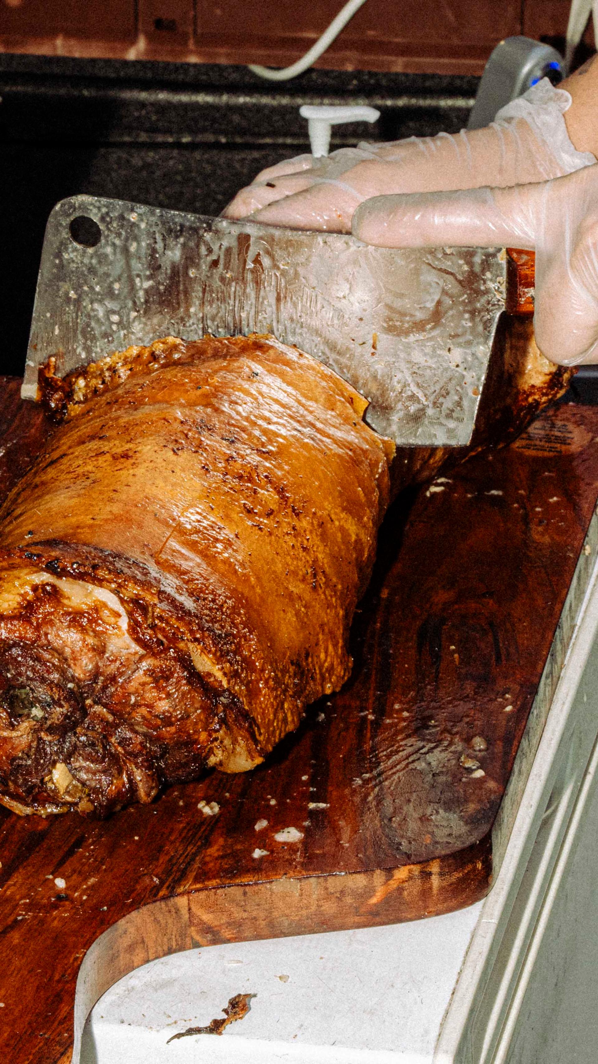 A vendor in gloves slices a large roasted meat piece on a wooden cutting board, with various kitchen tools and containers in the background.