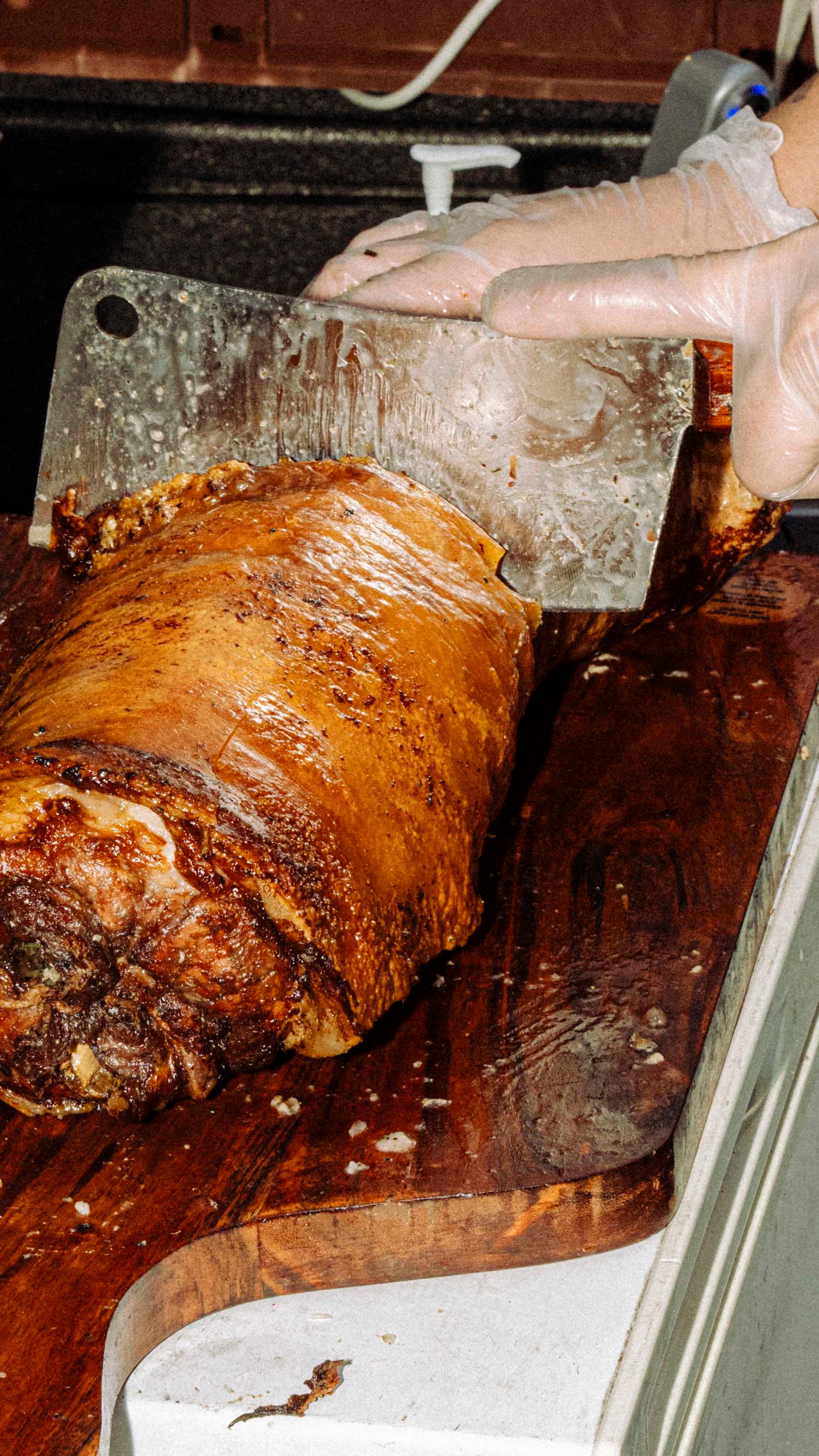 A vendor in gloves slices a large roasted meat piece on a wooden cutting board, with various kitchen tools and containers in the background.